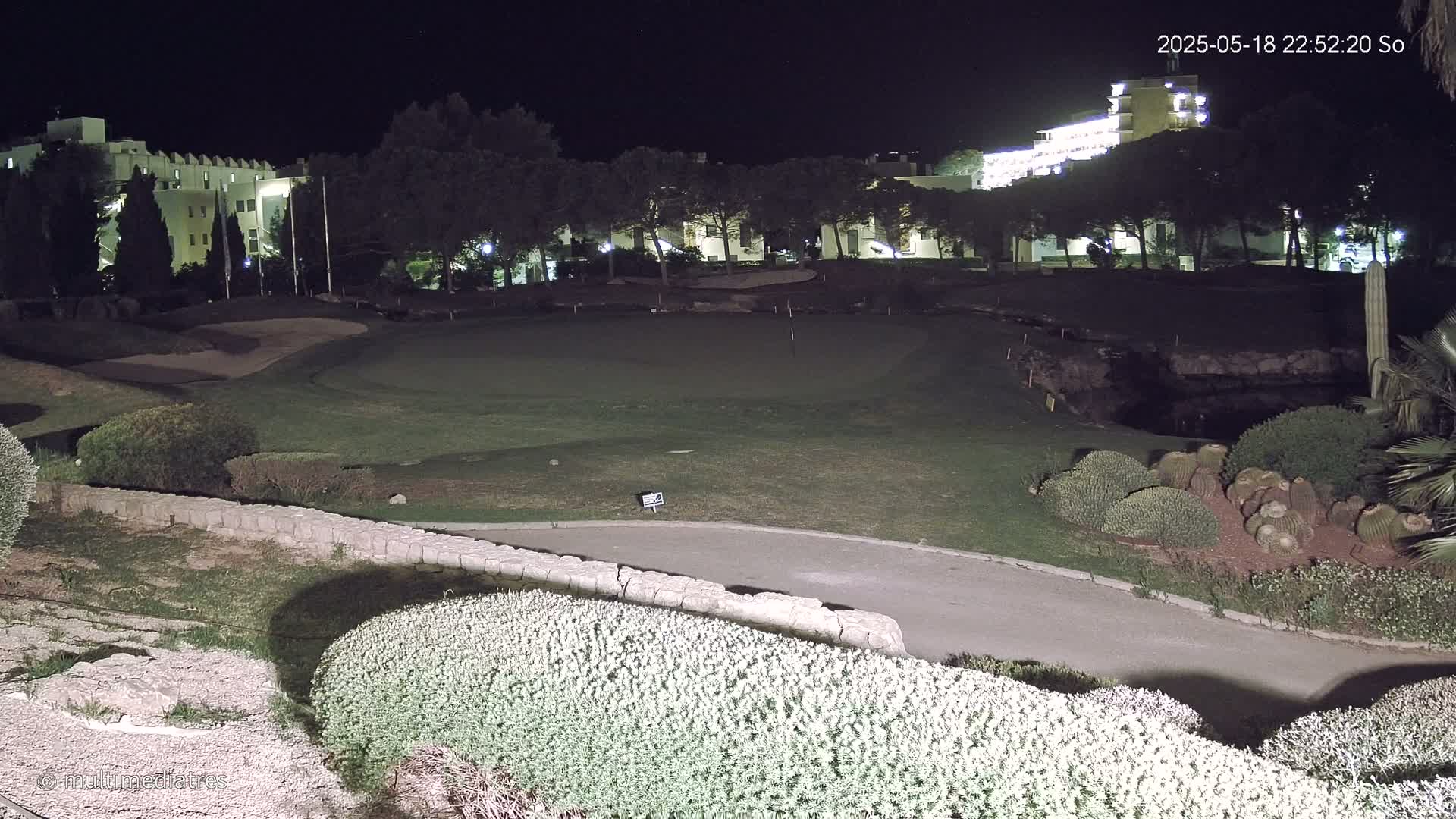 A nighttime view of a putting green on a golf course adjacent to a lit-up building complex.