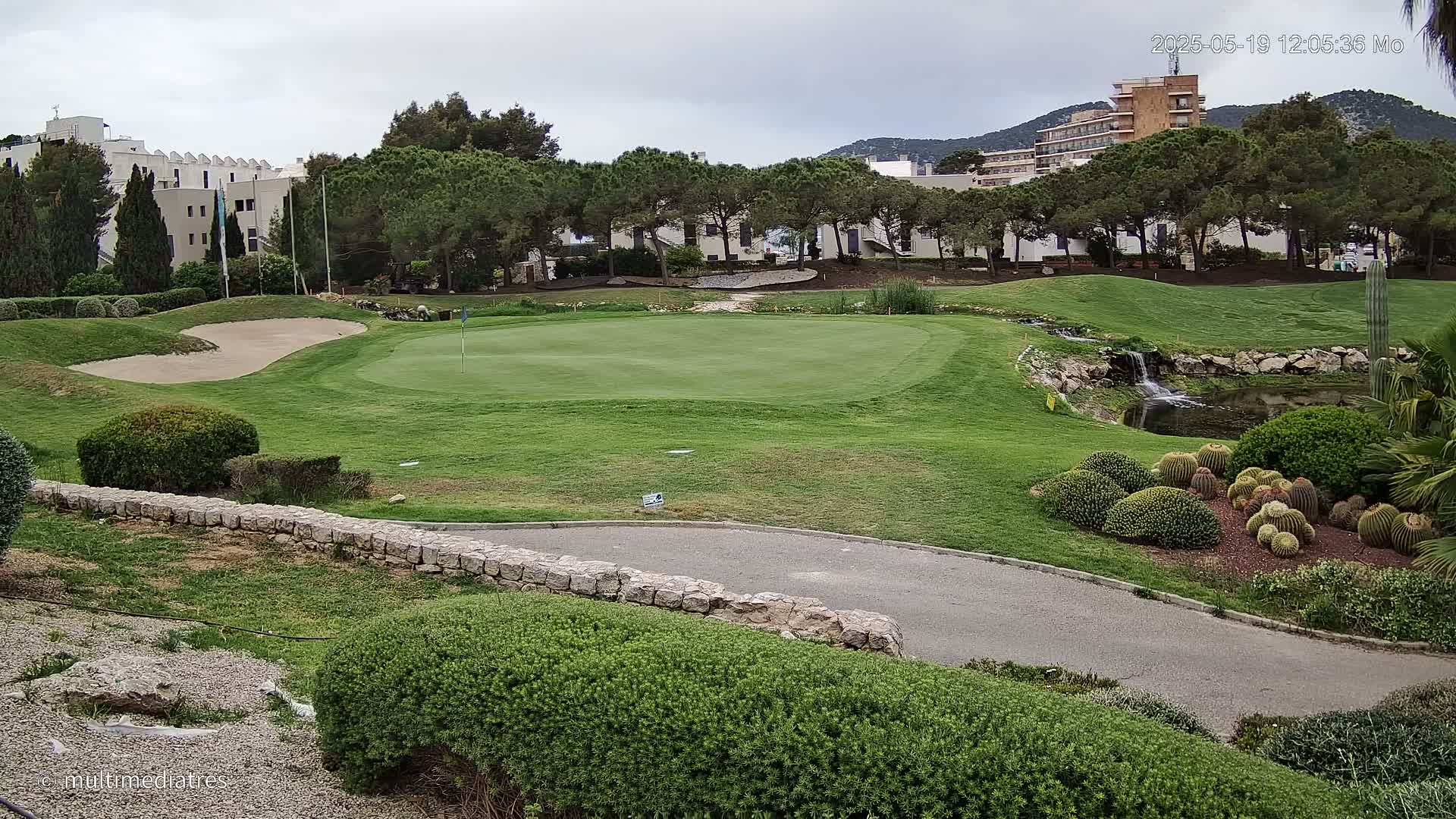 A golf course green with a sand trap, a small waterfall, and several cacti in the foreground, under an overcast sky.