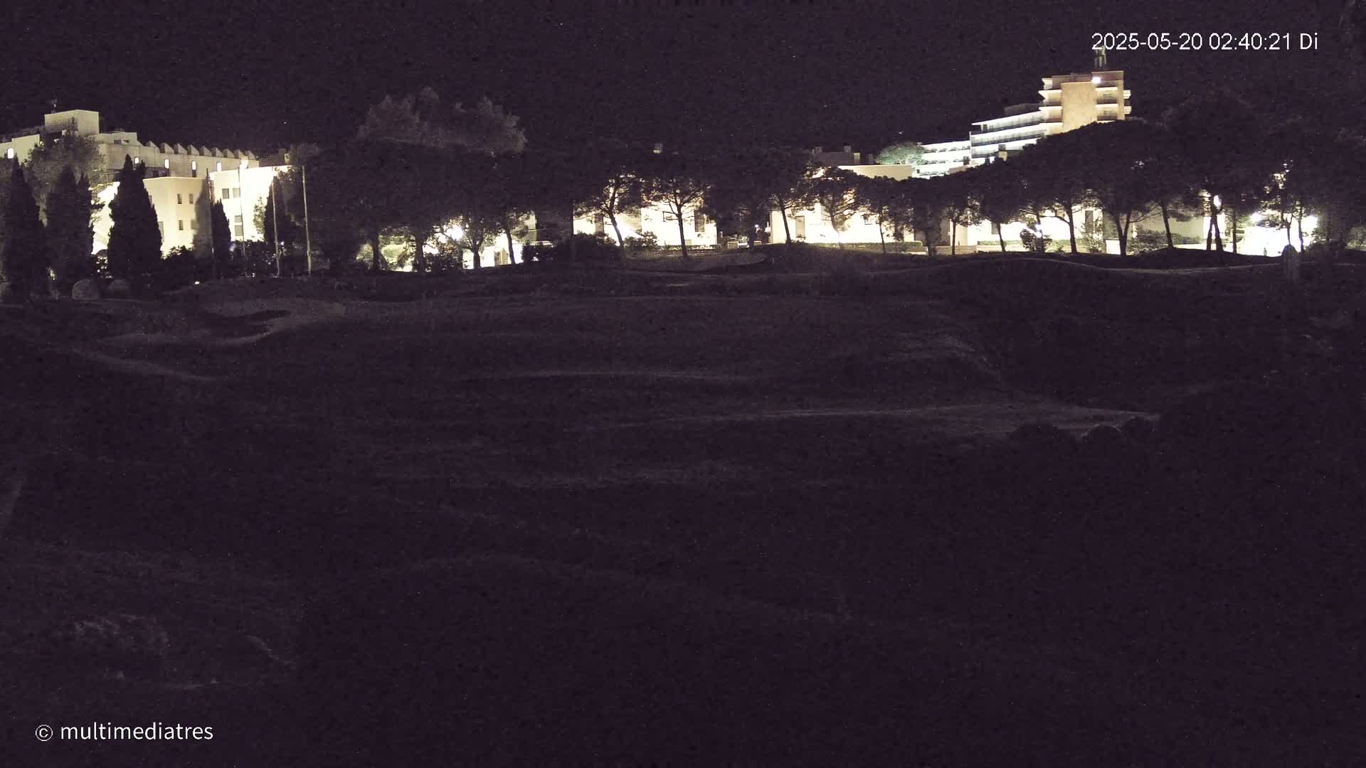 At night, illuminated buildings are visible behind a dark, uneven expanse of land, possibly a field or park, dotted with silhouetted trees.