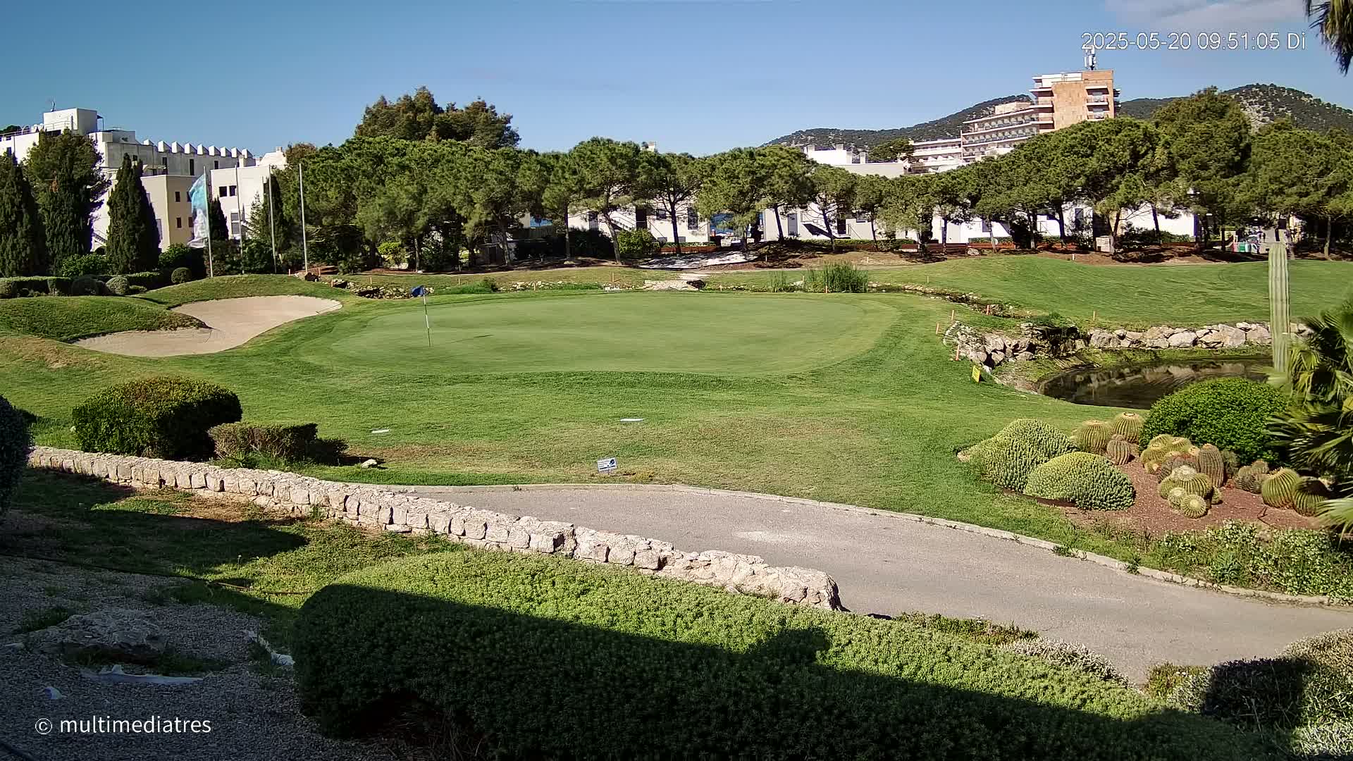 A golf course green with a sand trap and a small pond, surrounded by buildings and lush vegetation under a clear blue sky.