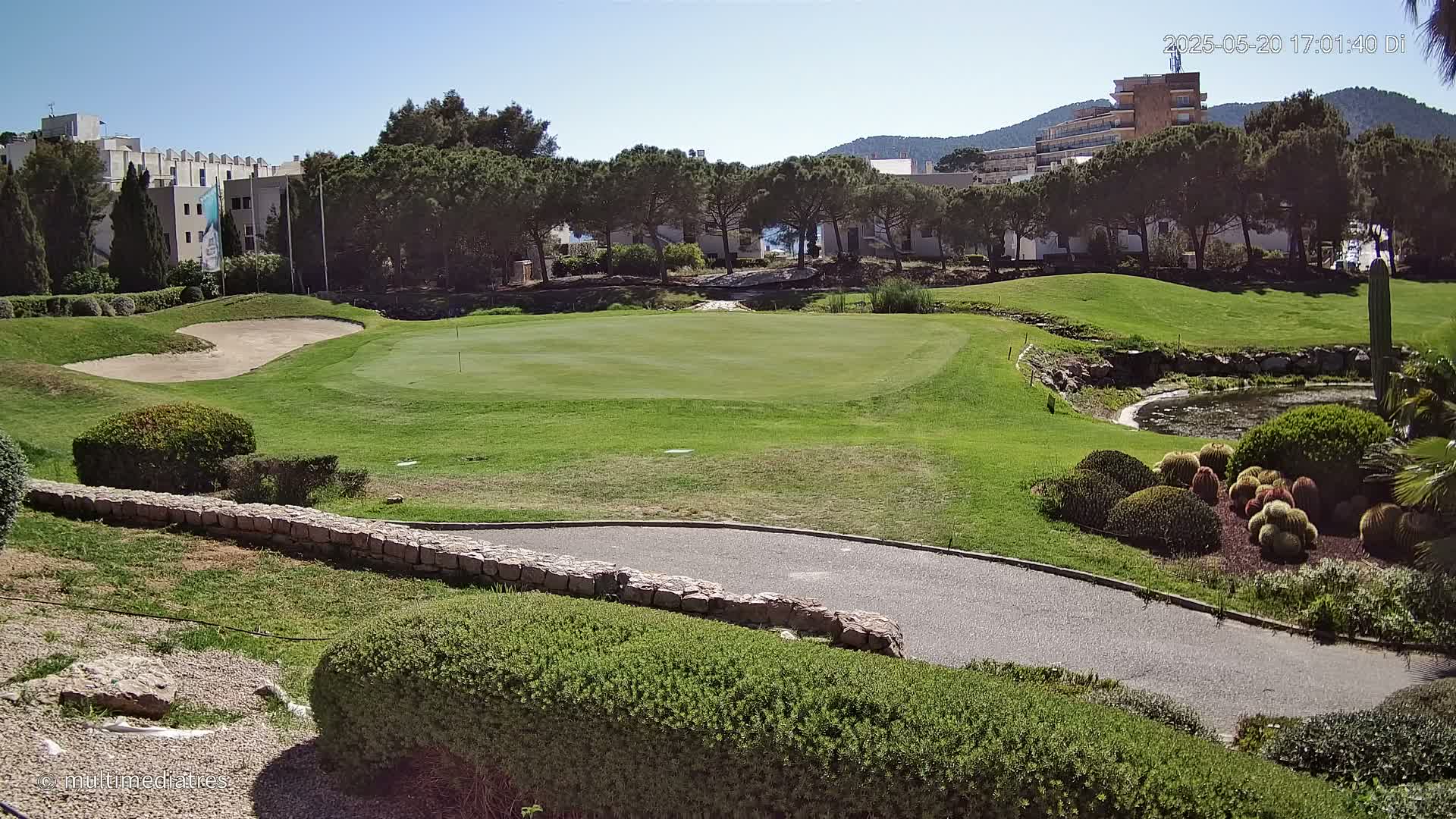 A golf course putting green with a sand trap, bordered by manicured shrubs and low stone walls, under a clear sunny sky.