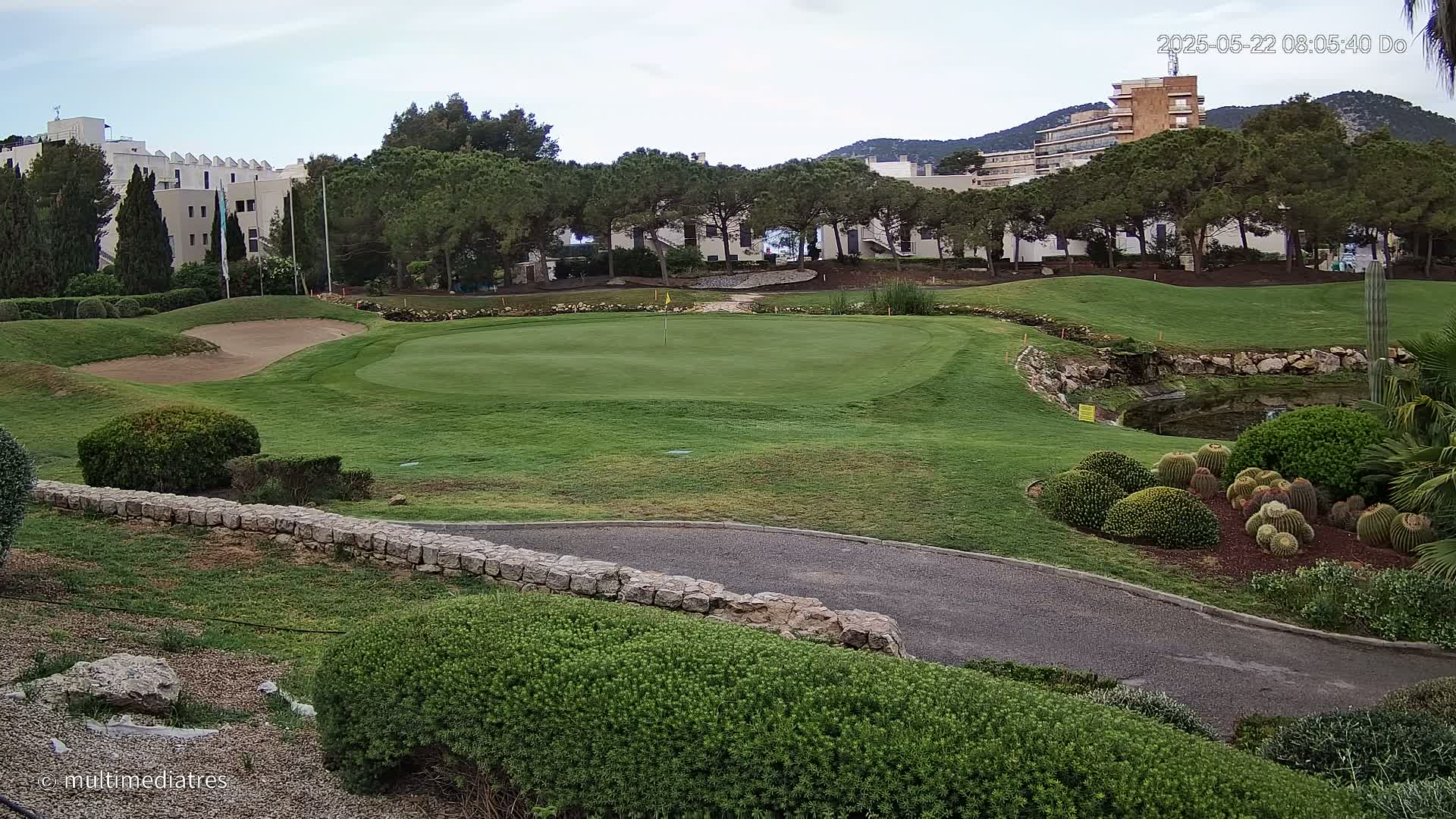 A golf course putting green with a sand trap and a landscaped area with cacti, under an overcast sky.