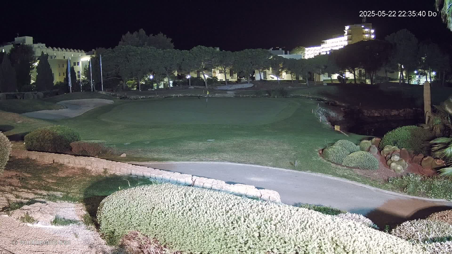 A nighttime view of a golf course green, bordered by shrubs and low stone walls, with illuminated buildings in the background under a clear, dark sky.