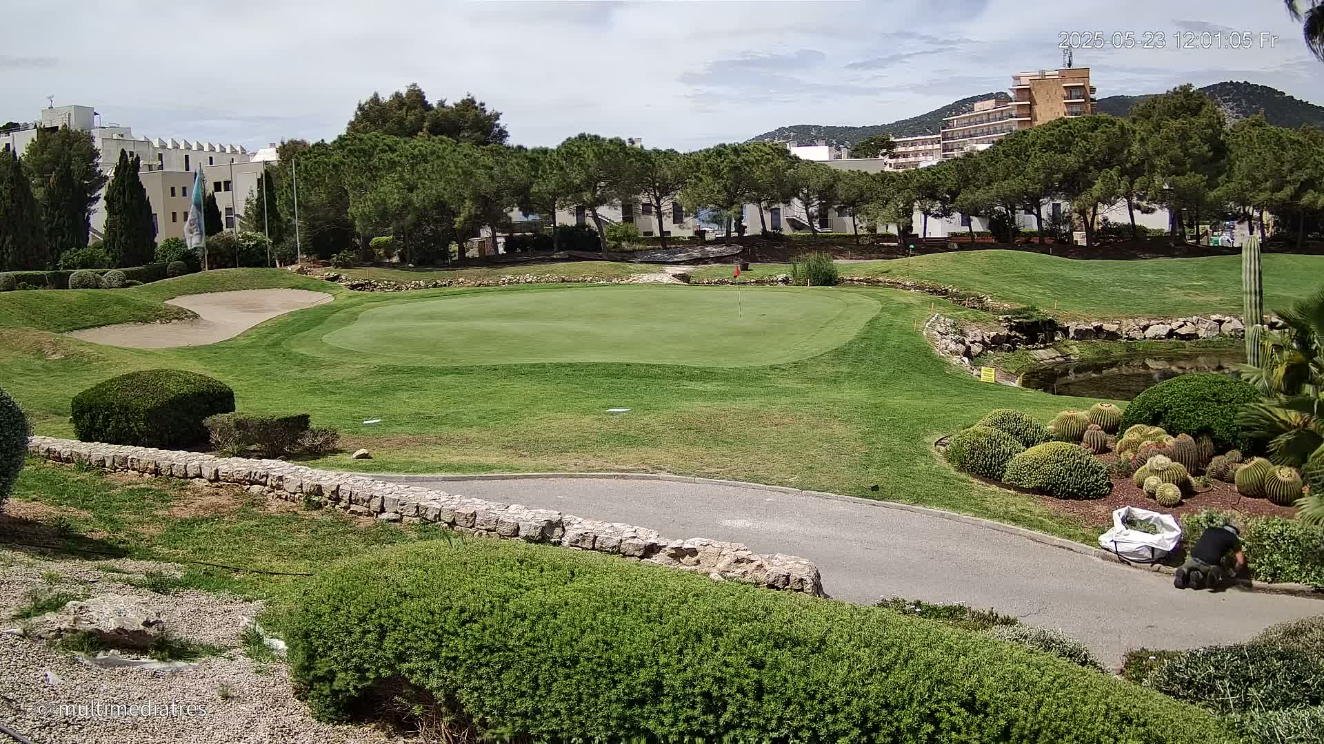 A golf course green with a sand trap, landscaping featuring cacti, and buildings in the background under partly cloudy skies.