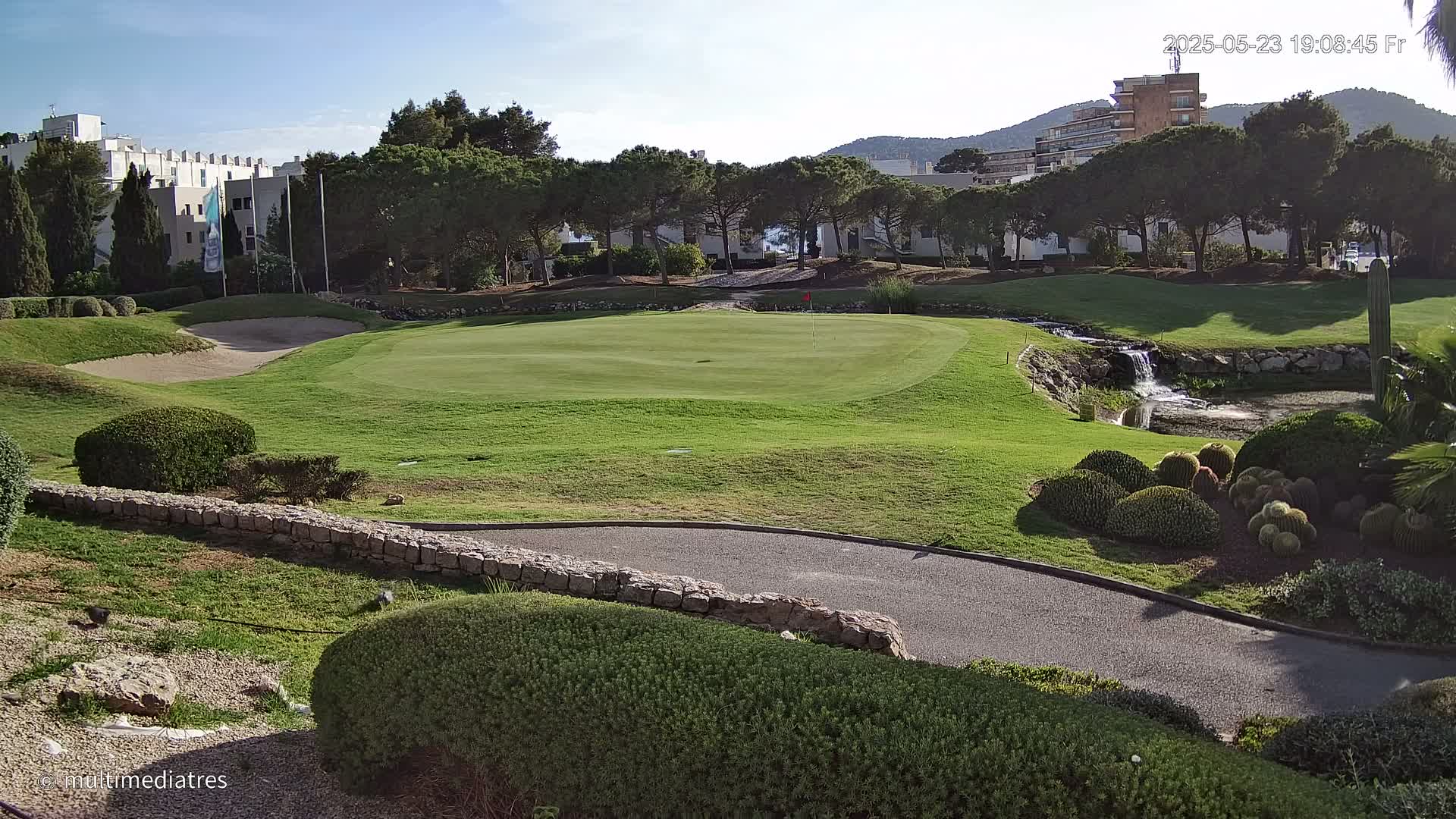 A putting green on a golf course is situated near a small waterfall and pond, with buildings and trees visible in the background under a clear sky.