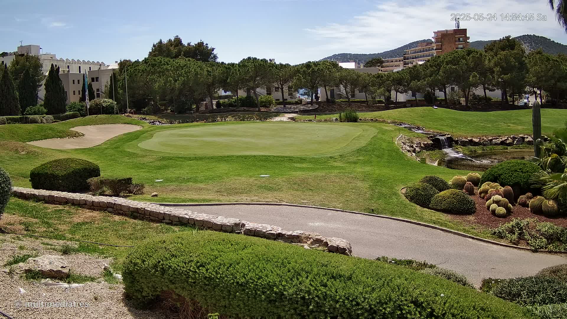 A golf course green with a sand trap, a small waterfall, and manicured landscaping under a sunny sky.