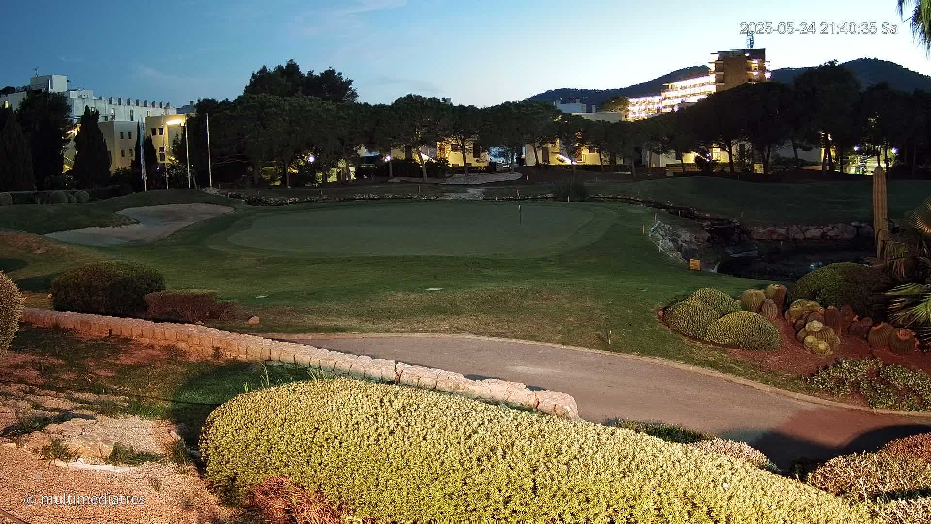 A golf course green is illuminated at night, with buildings and trees visible in the background under a twilight sky.