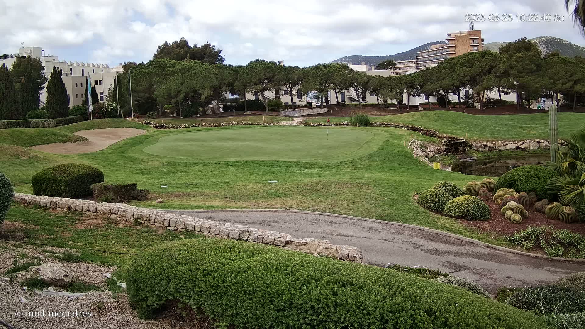 A golf course green, sand trap, and cart path are surrounded by landscaping, trees, and buildings under a partly cloudy sky.