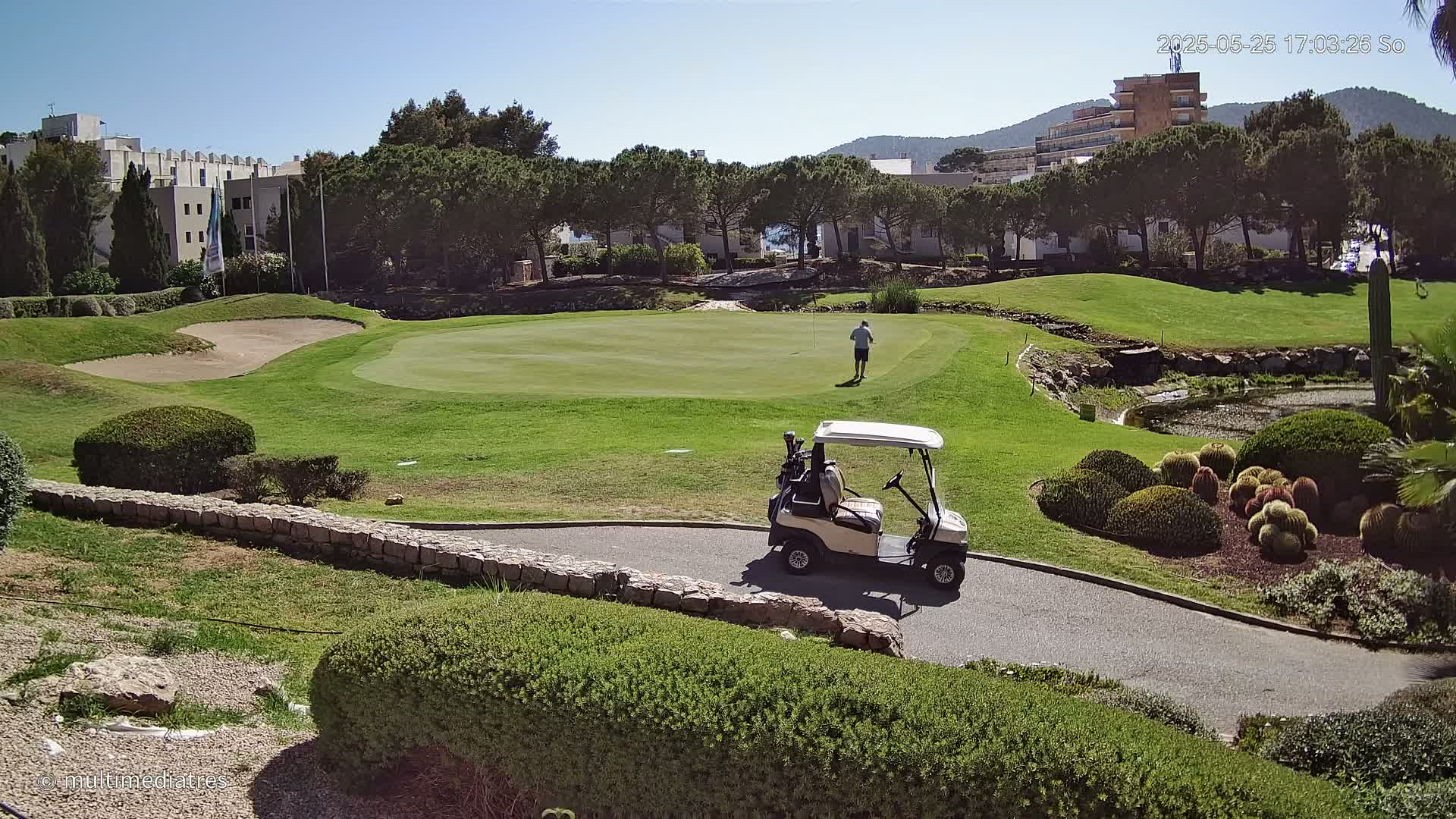 A golfer walks toward a green on a sunny day, a golf cart parked nearby on a paved path beside a stone wall, with manicured landscaping and buildings visible in the background.