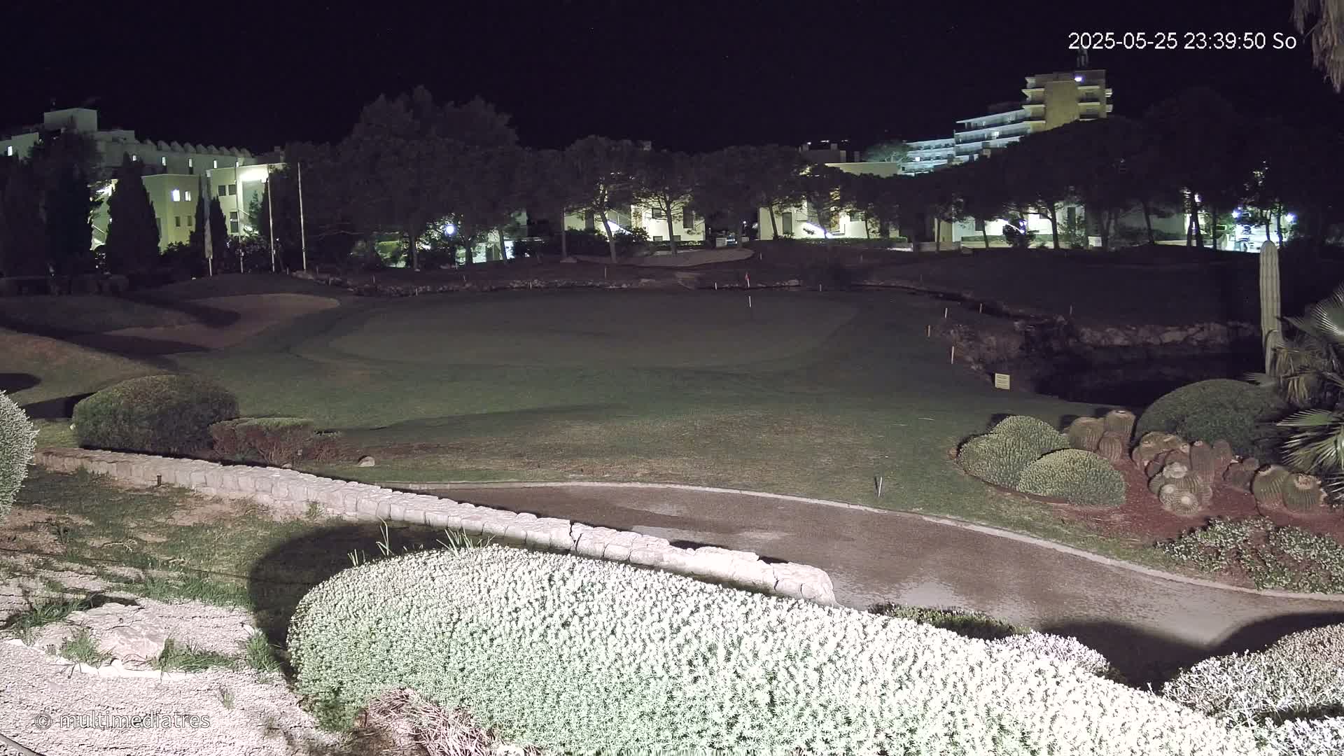 A nighttime view of a golf course, illuminated by artificial light, with a building visible in the background.