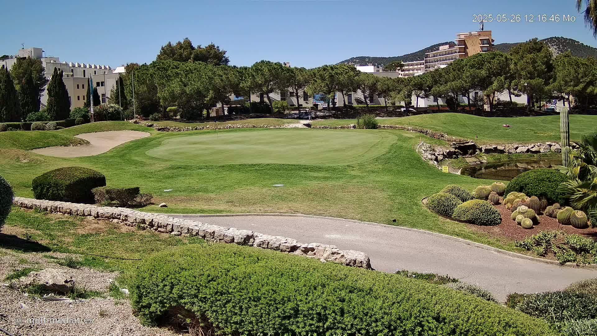 A golf course putting green is visible, surrounded by manicured lawns, shrubs, and a low stone wall, under a bright sunny sky.