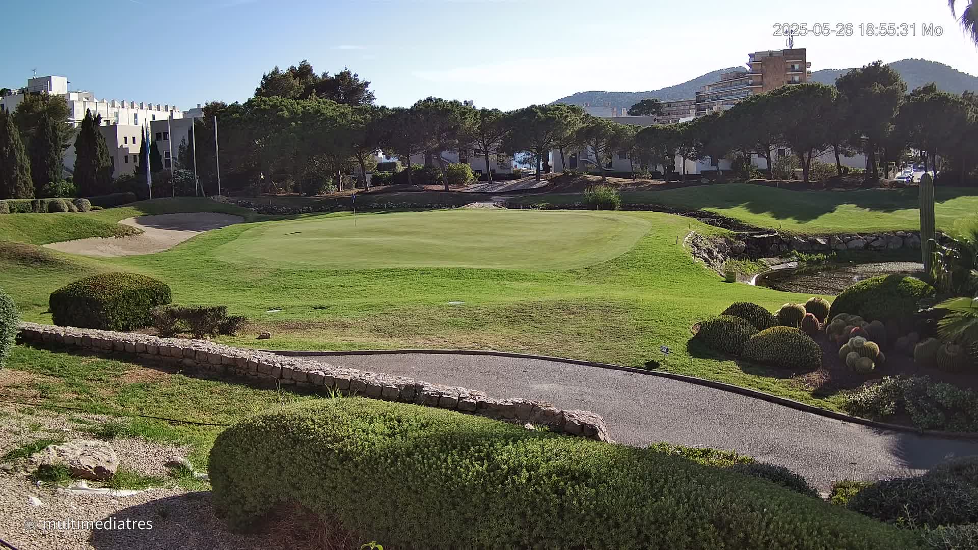 A golf course green, sand trap, and paved path are visible under a sunny sky, with buildings and trees in the background.