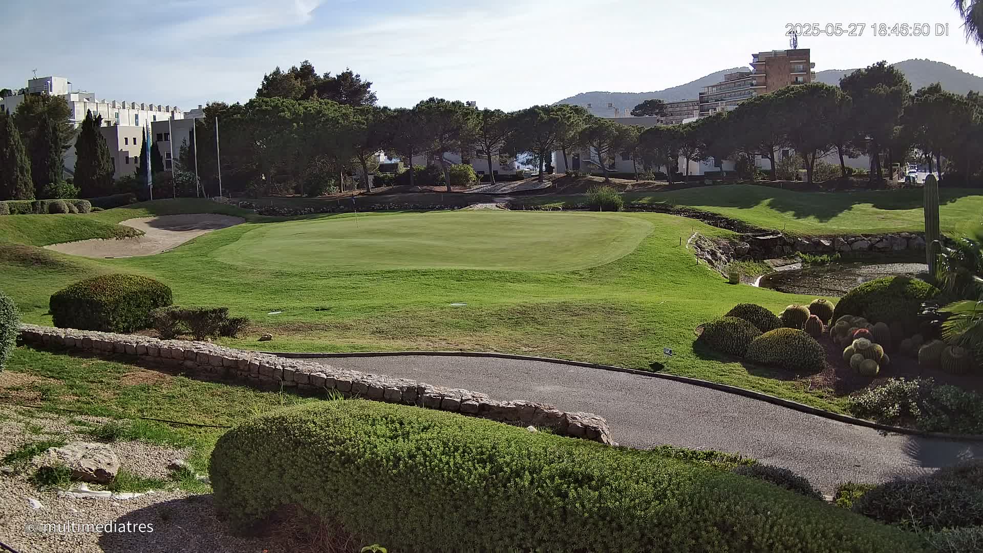 A golf course green with a sand trap and a small pond, surrounded by low shrubs, a stone wall, and buildings in the background under a clear, sunny sky.