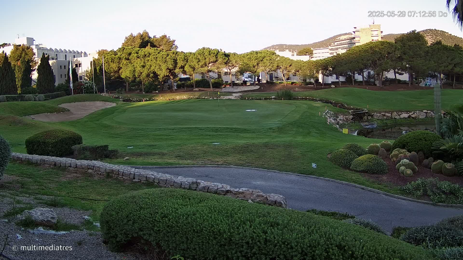 A golf course green, bordered by manicured landscaping and buildings, is bathed in the soft light of early morning or late afternoon.