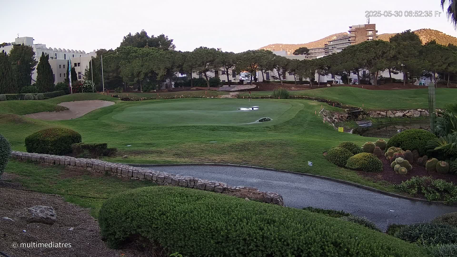 A golf course green with a sand trap and a low stone wall, bordered by lush shrubs and cacti, is situated near buildings under a mostly clear sky.