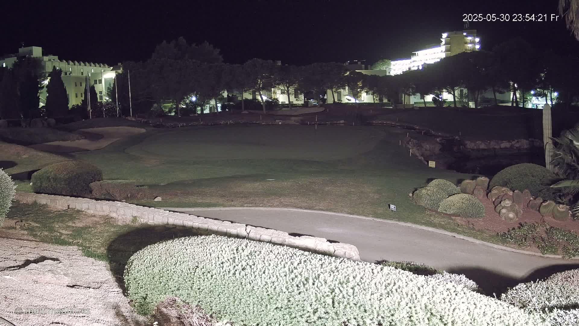 A nighttime view of a golf course putting green, bordered by shrubs and a low stone wall, with illuminated buildings in the background under a clear, dark sky.
