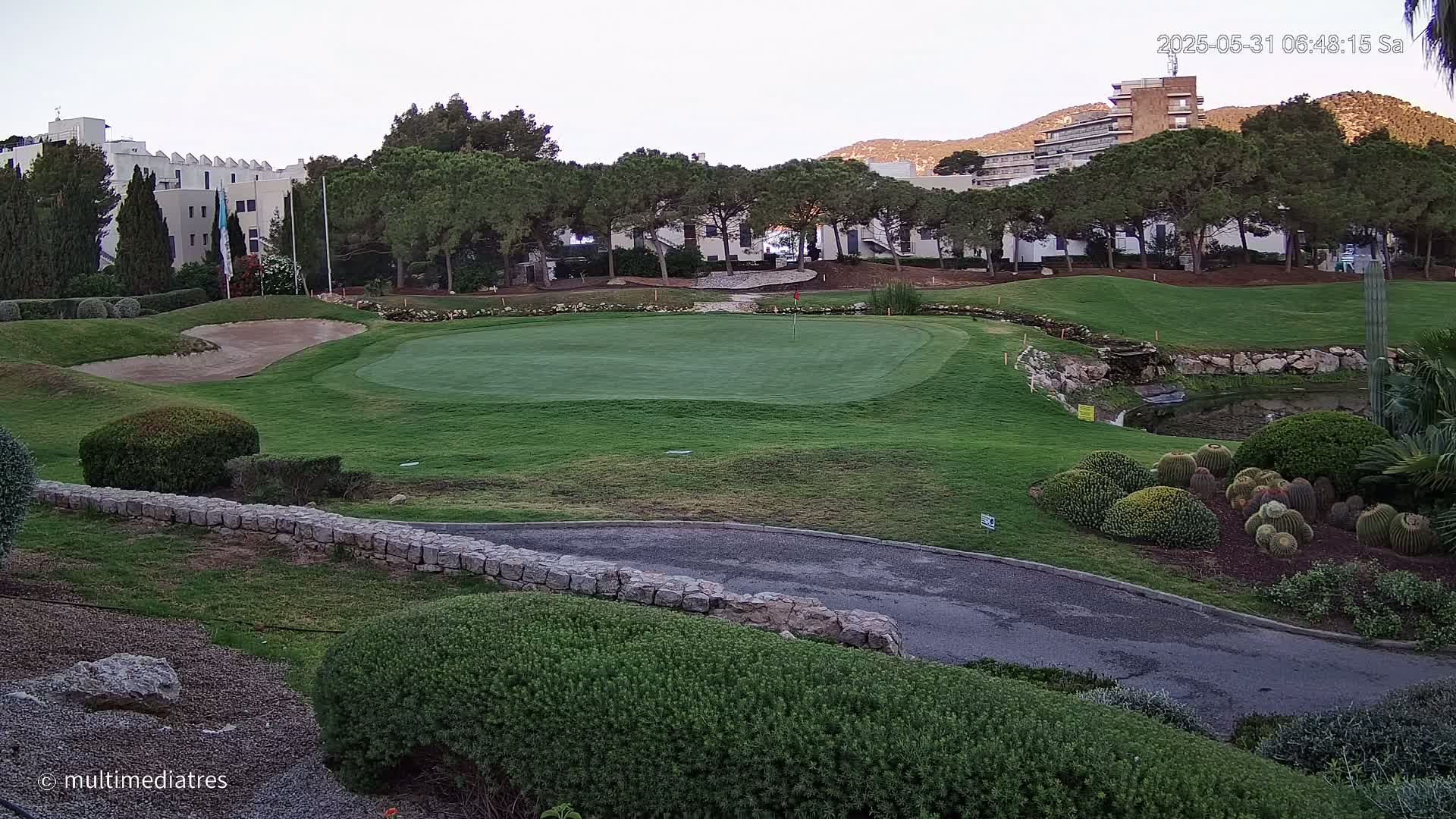 A golf course green with sand traps, a small pond, and manicured landscaping, under a partly cloudy sky.