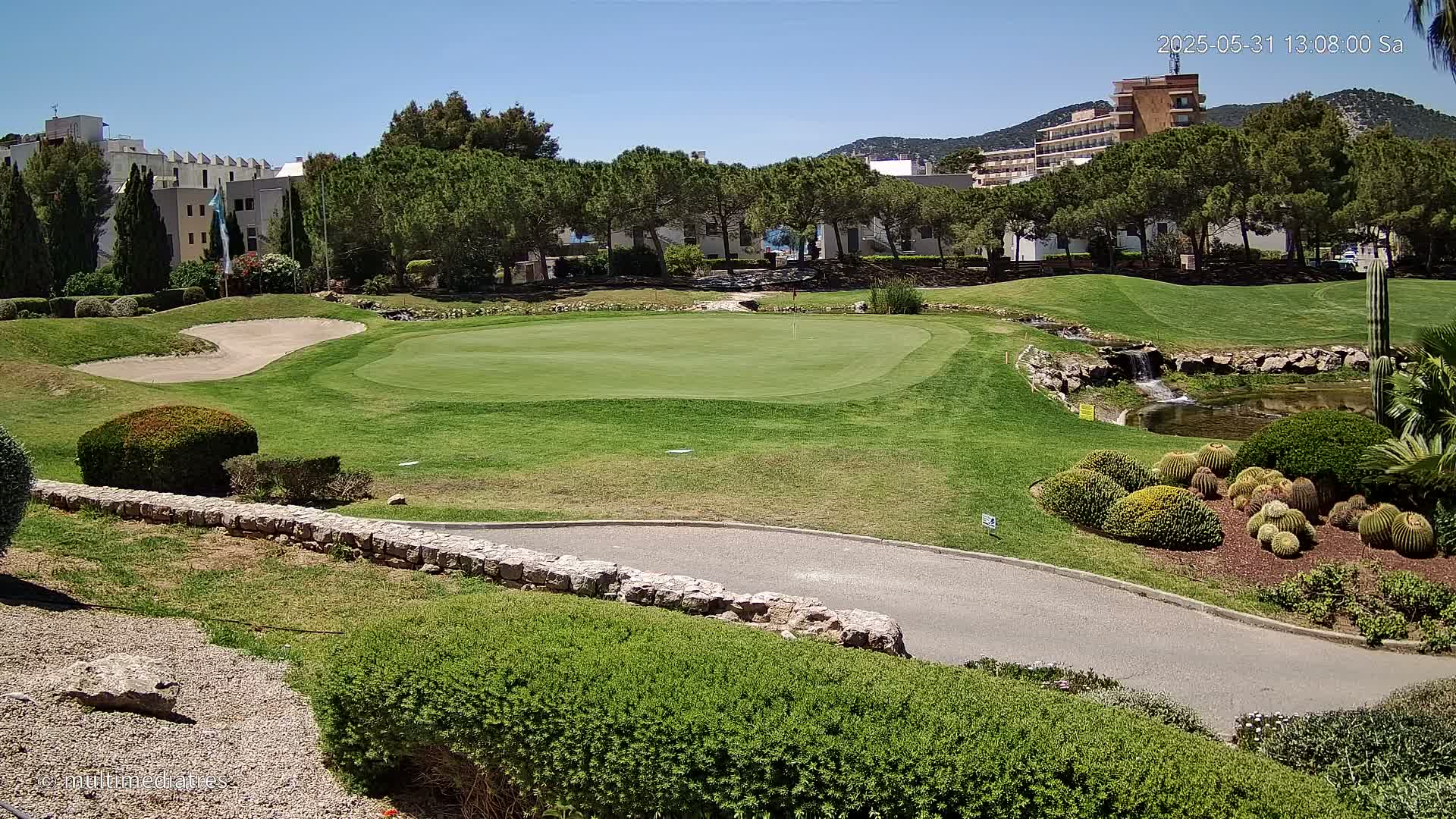 A putting green on a golf course is surrounded by manicured lawns, shrubs, and a small waterfall under a clear, sunny sky.