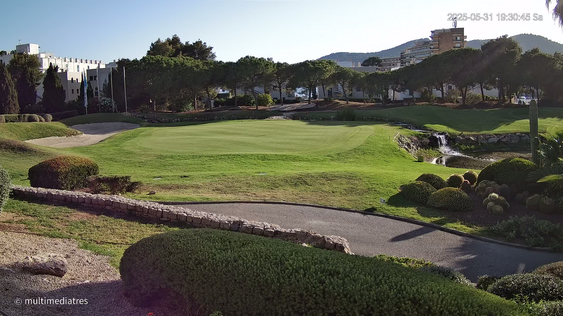 A golf course green with a small waterfall and pond, surrounded by manicured landscaping and buildings in the background under a clear, sunny sky.