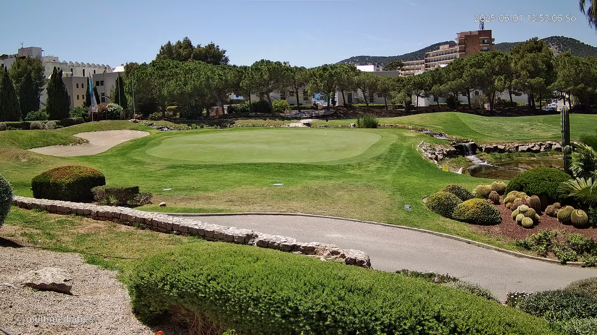 A golf course putting green with a sand trap, a small waterfall, and manicured landscaping, under a clear sunny sky.