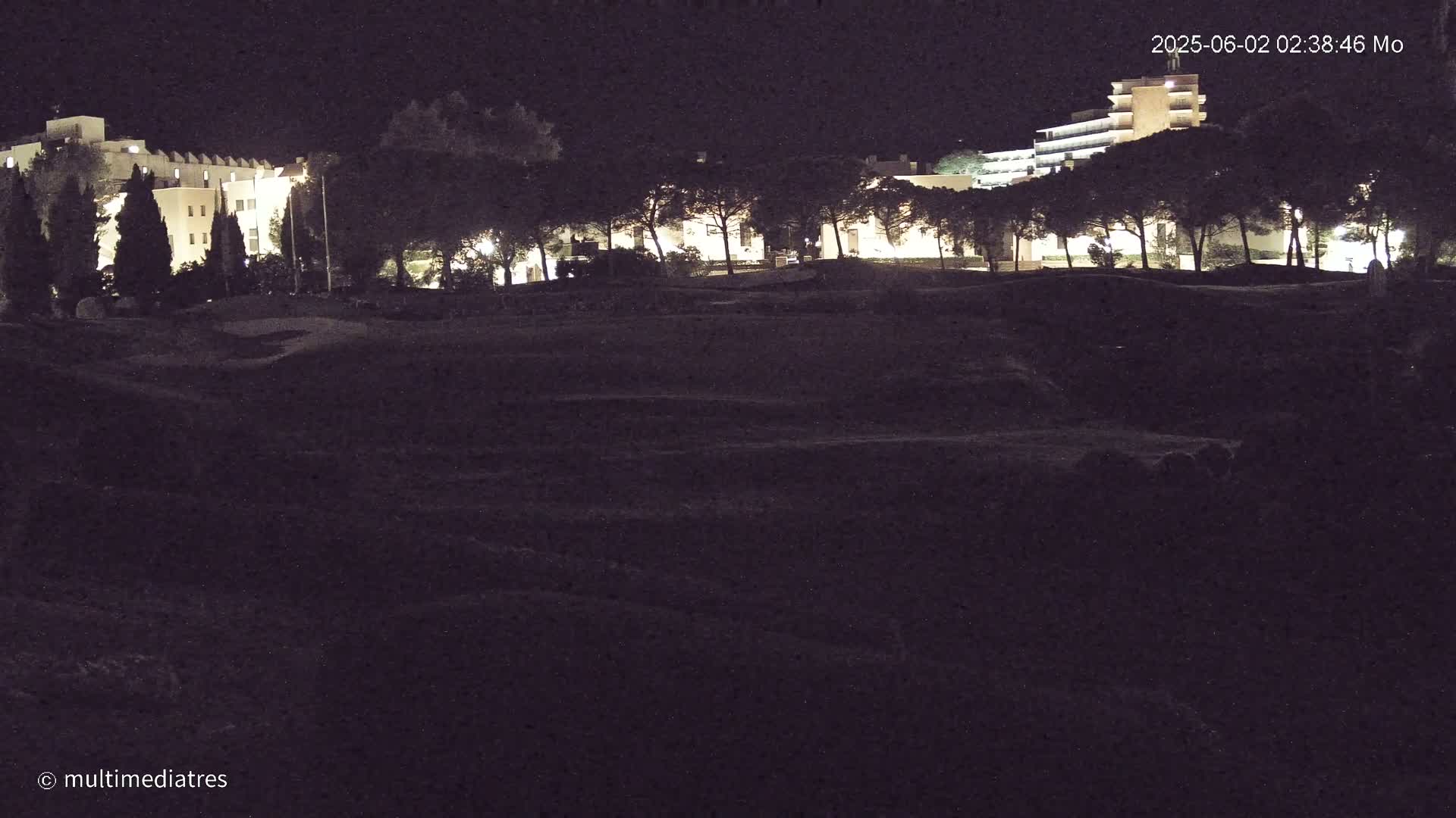 At night, illuminated buildings are visible beyond a dark, grassy expanse.