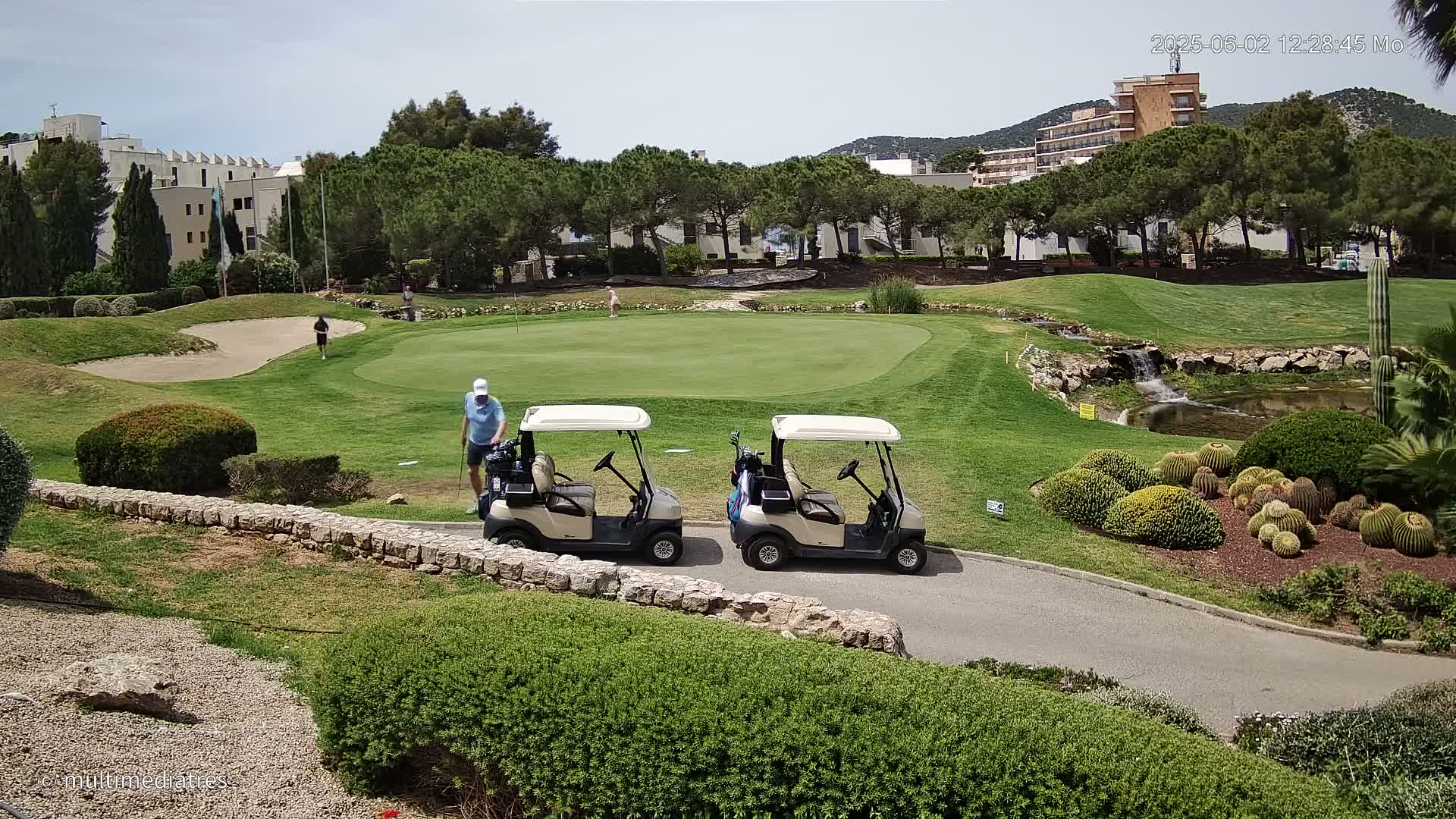 Two golf carts are parked near a putting green on a sunny day, with a small waterfall and cacti visible in the foreground.