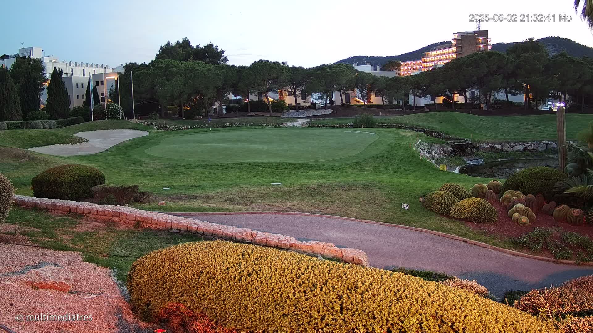 A golf course green, bordered by landscaping and a low stone wall, sits in front of a building complex at dusk.