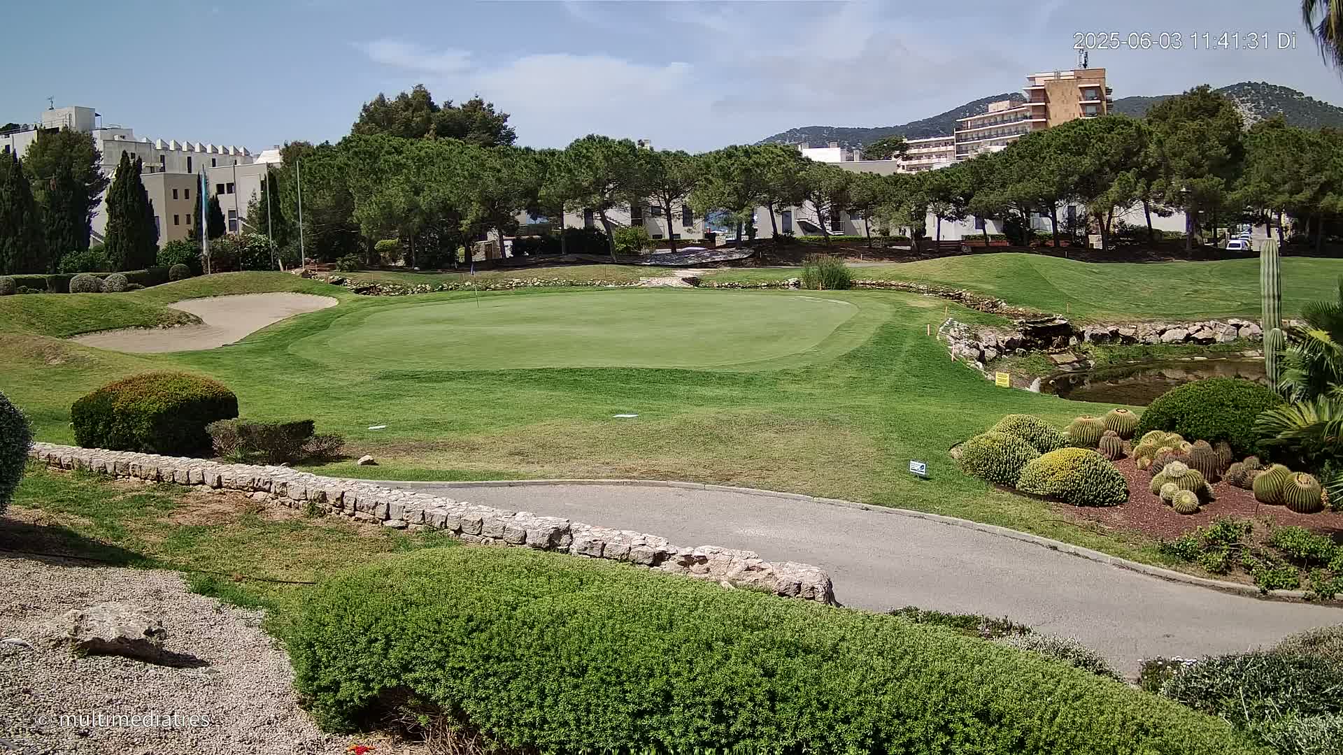 A golf course putting green with a sand trap and buildings in the background under sunny skies.