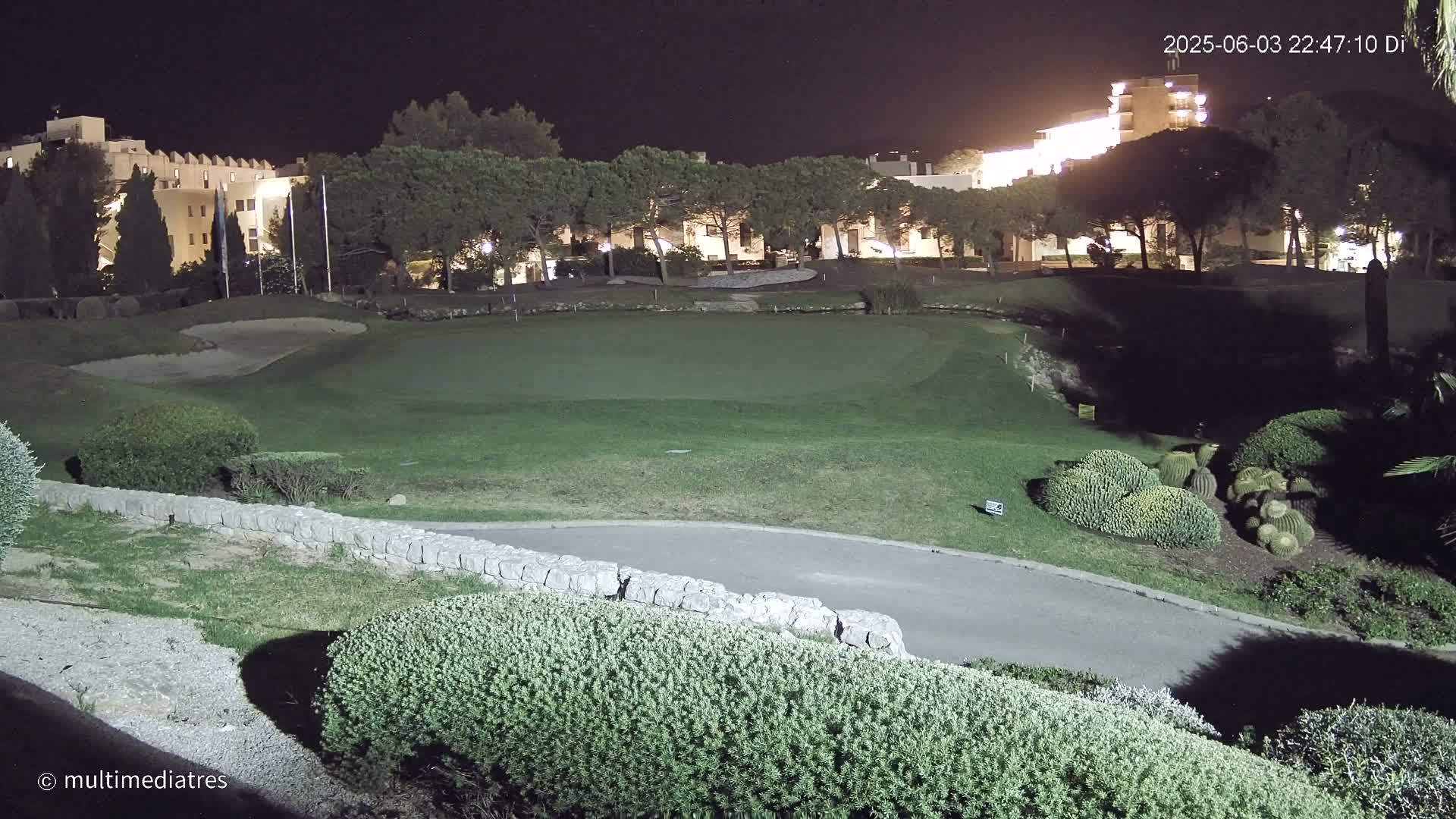 A well-lit golf course at night, with a putting green and surrounding landscaping, is situated near a building complex.
