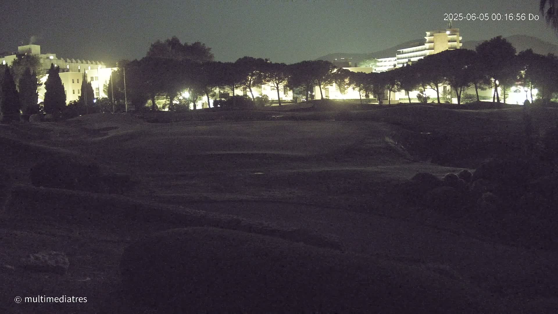 A nighttime view of a grassy area with buildings and trees in the background under a dark sky.