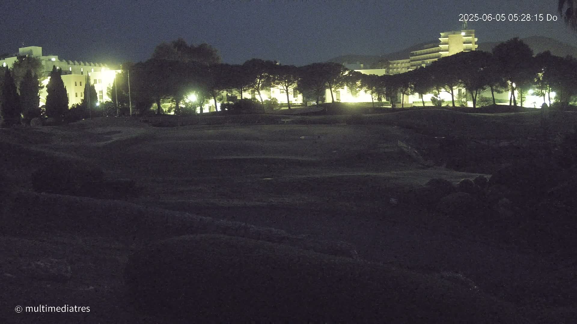 At night, a dark, uneven expanse of land sits in front of a row of illuminated buildings and trees.