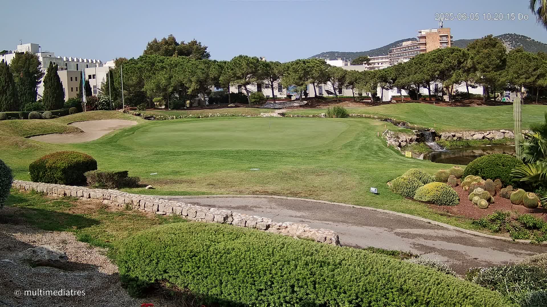 A golf course green with a sand trap, a small waterfall, and cacti, all under a clear sunny sky.