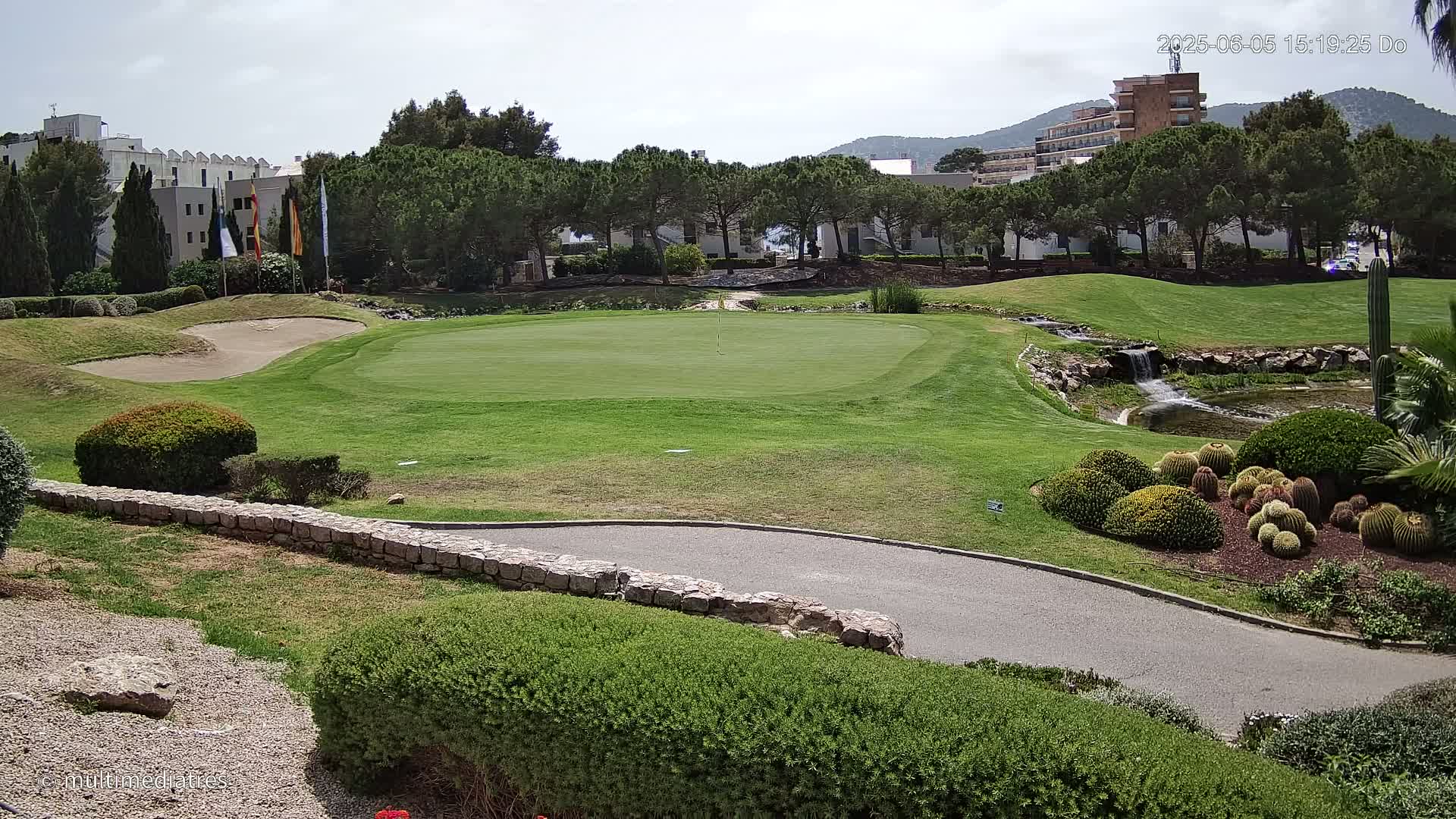 A golf course green with a sand trap, a small waterfall, and a variety of shrubs and cacti under a partly sunny sky.