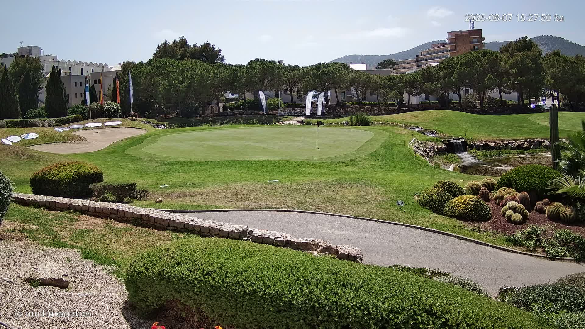 A sunny view of a golf course green, sand trap, and manicured landscaping featuring a small waterfall and cacti, with buildings visible in the background.