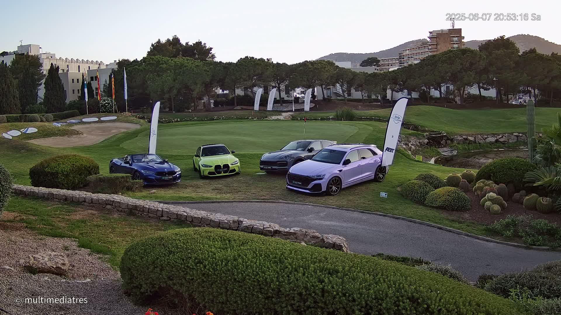 Four luxury cars are parked on a putting green at a golf course under a clear evening sky.