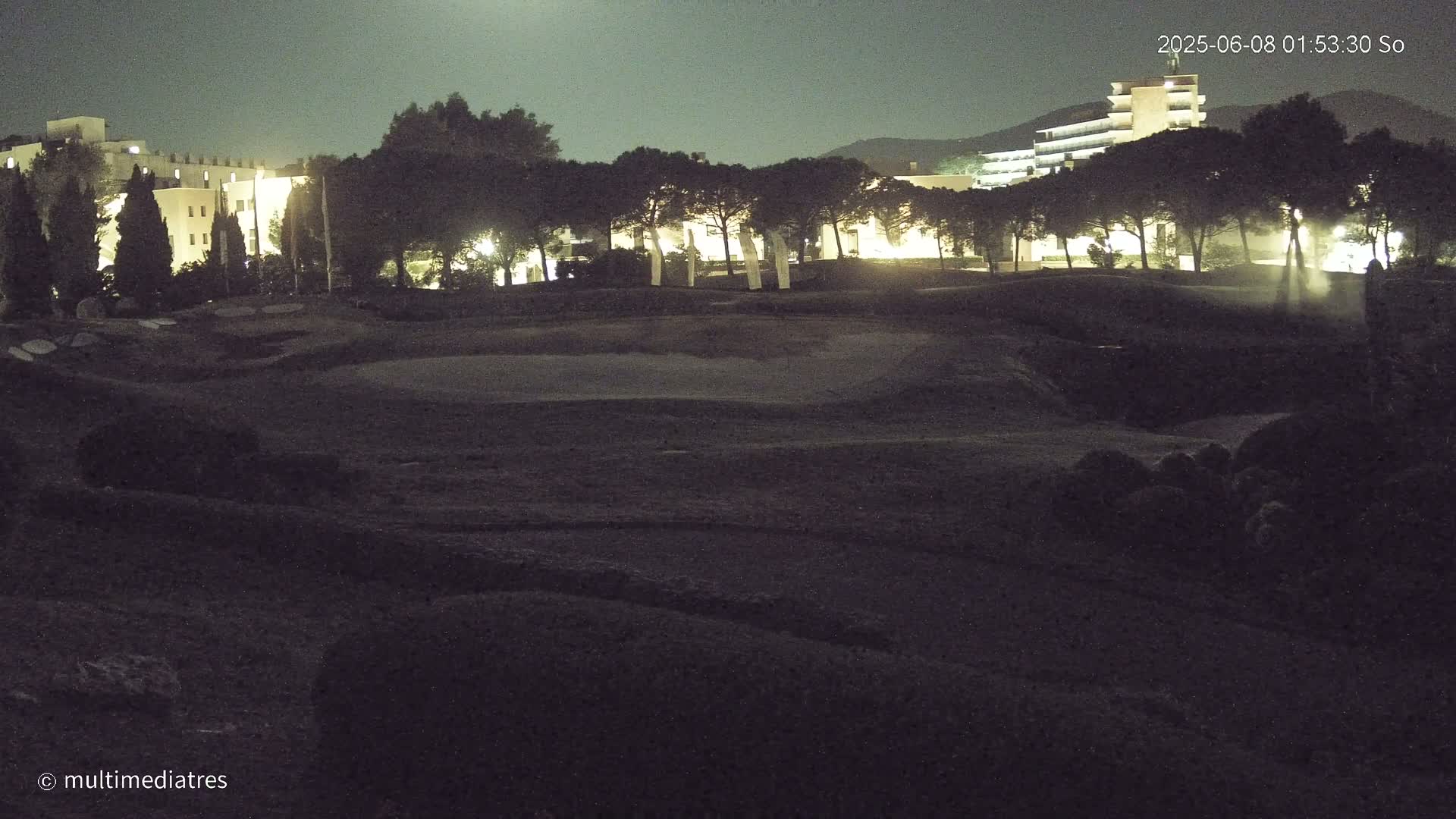 A nighttime view of a golf course with illuminated buildings and trees in the background under a dark sky.