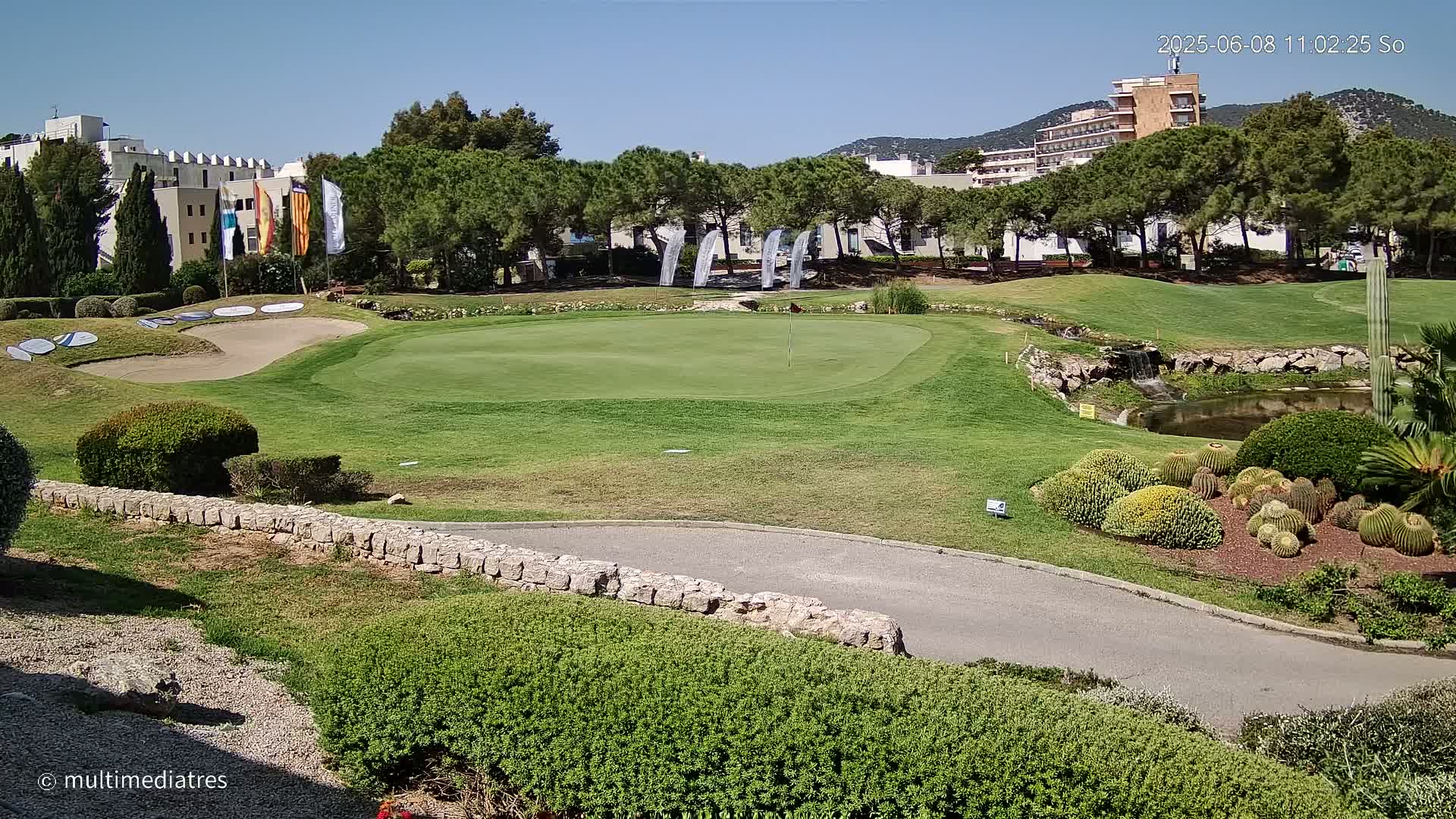 A golf course putting green with sand traps, a small pond, and cactus plants is visible under a clear, sunny sky, with buildings and trees in the background.