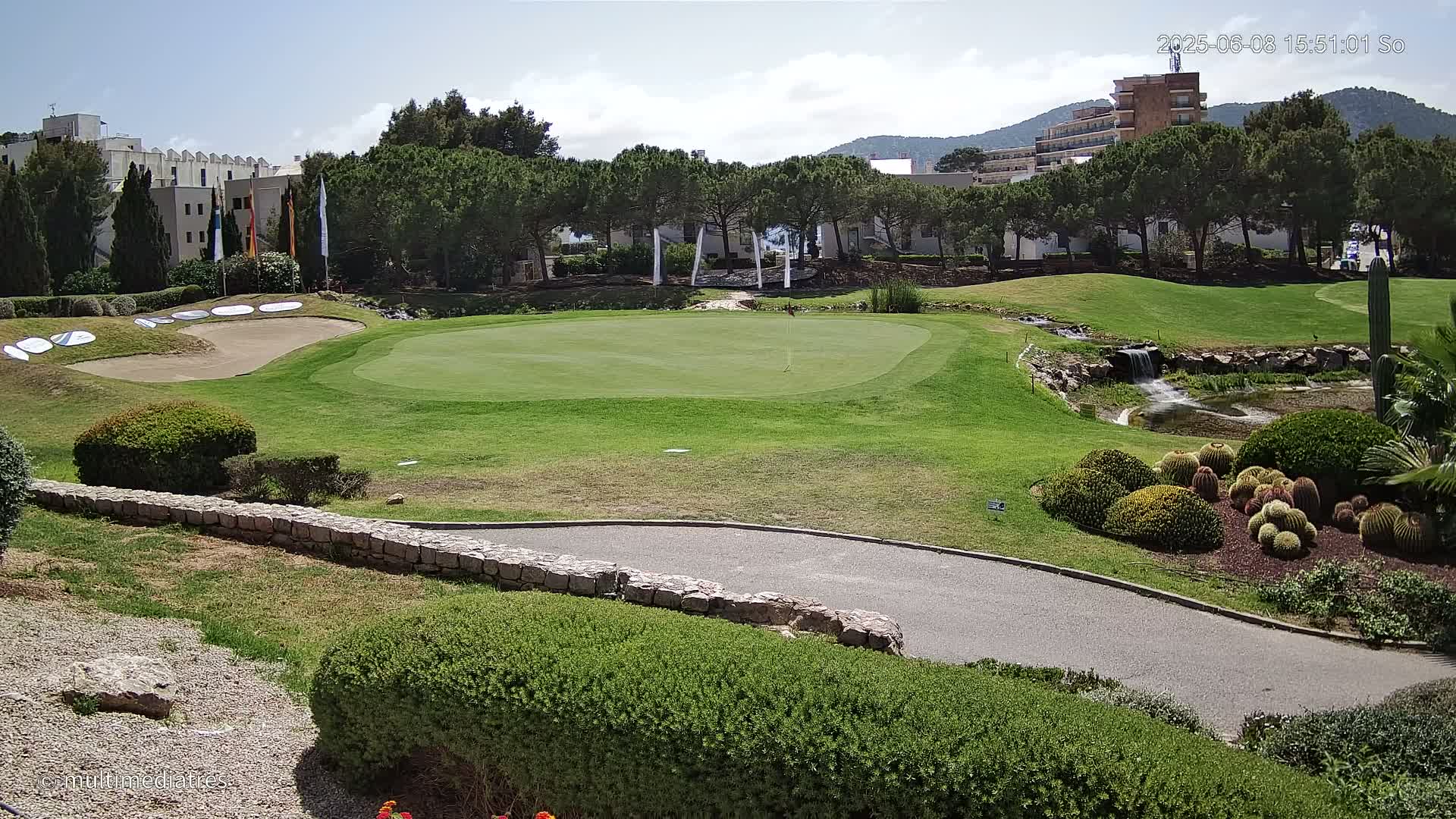 A golf course putting green with sand traps, a small waterfall feature, and cactus plants, all under a sunny sky.