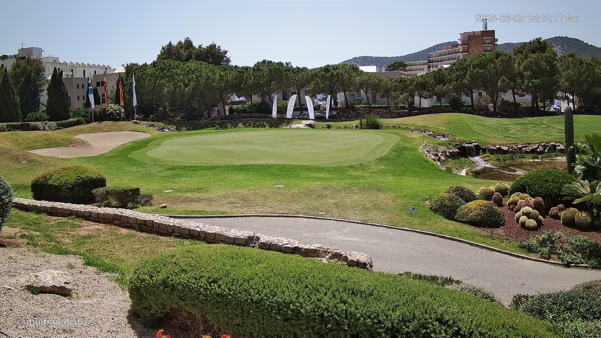 A putting green on a sunny day, bordered by landscaping, a low stone wall, and a small waterfall feature, with buildings and trees in the background.