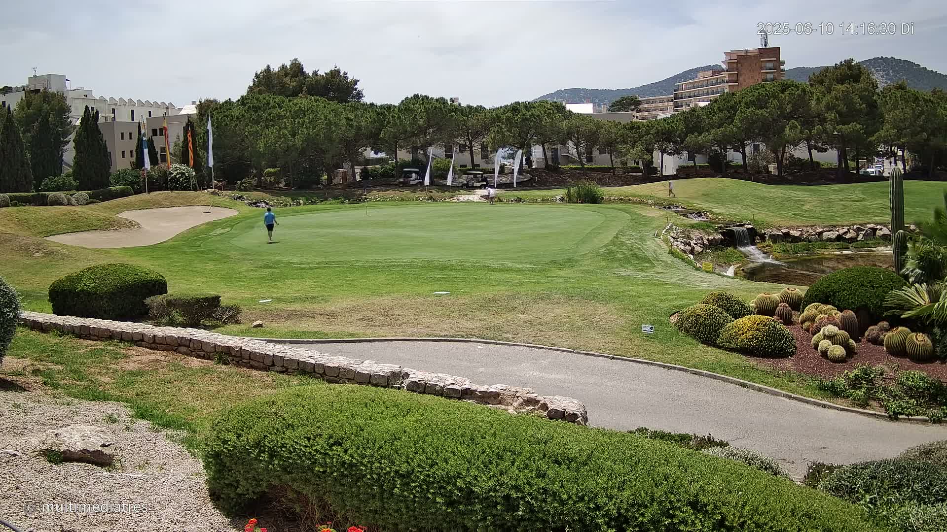 A golfer walks across a putting green on a sunny day, with a sand trap, a small waterfall, and resort buildings in the background.