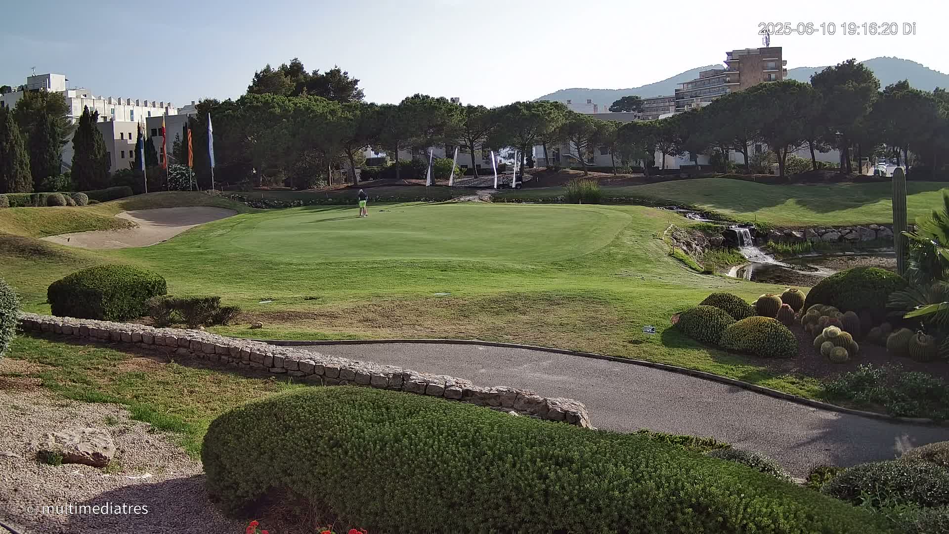 A golfer stands on a green putting surface of a golf course with a small waterfall and manicured landscaping in the sunny daytime.