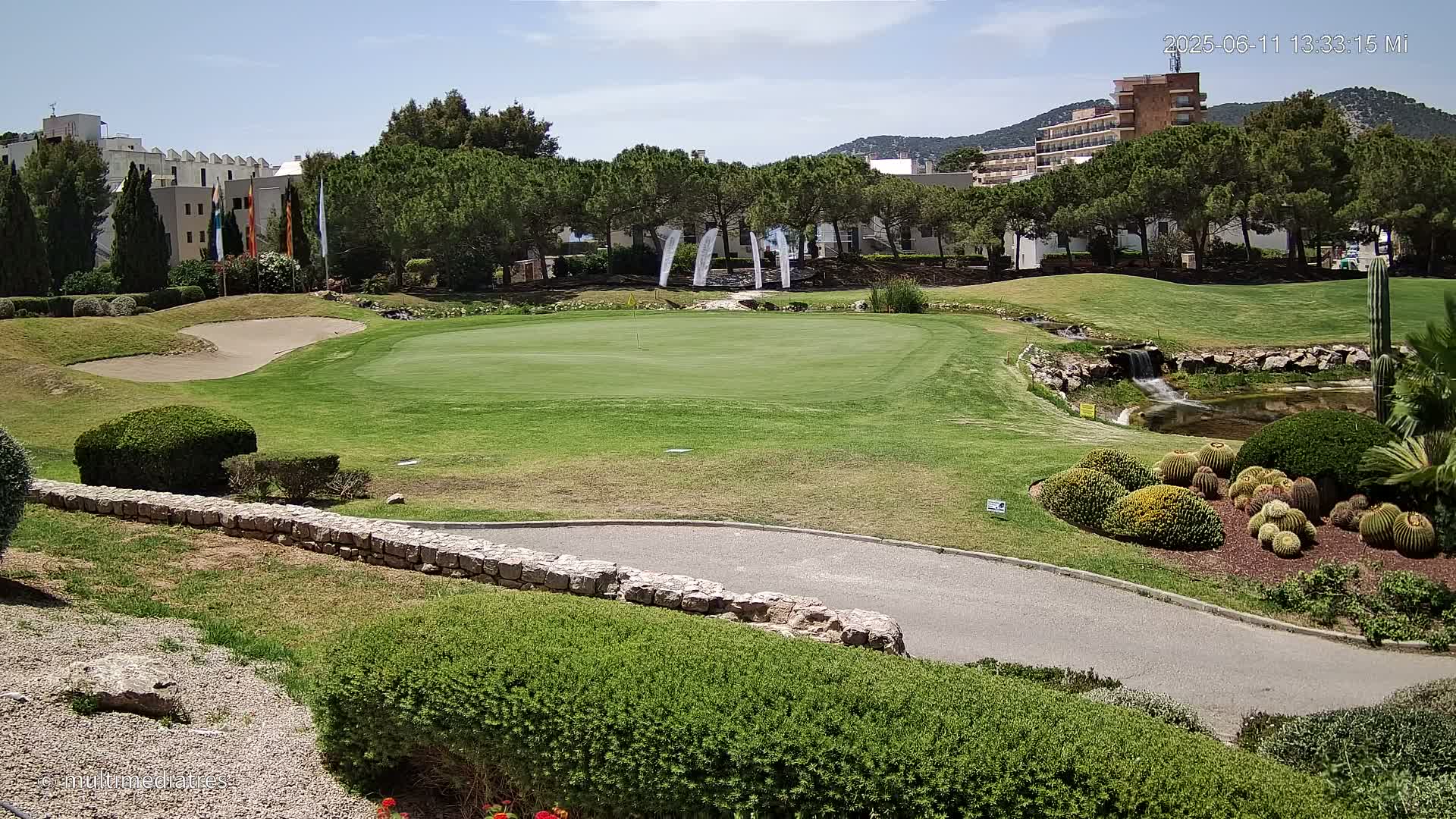 A golf course green with a sand trap, water feature, and several buildings in the background under a sunny sky.