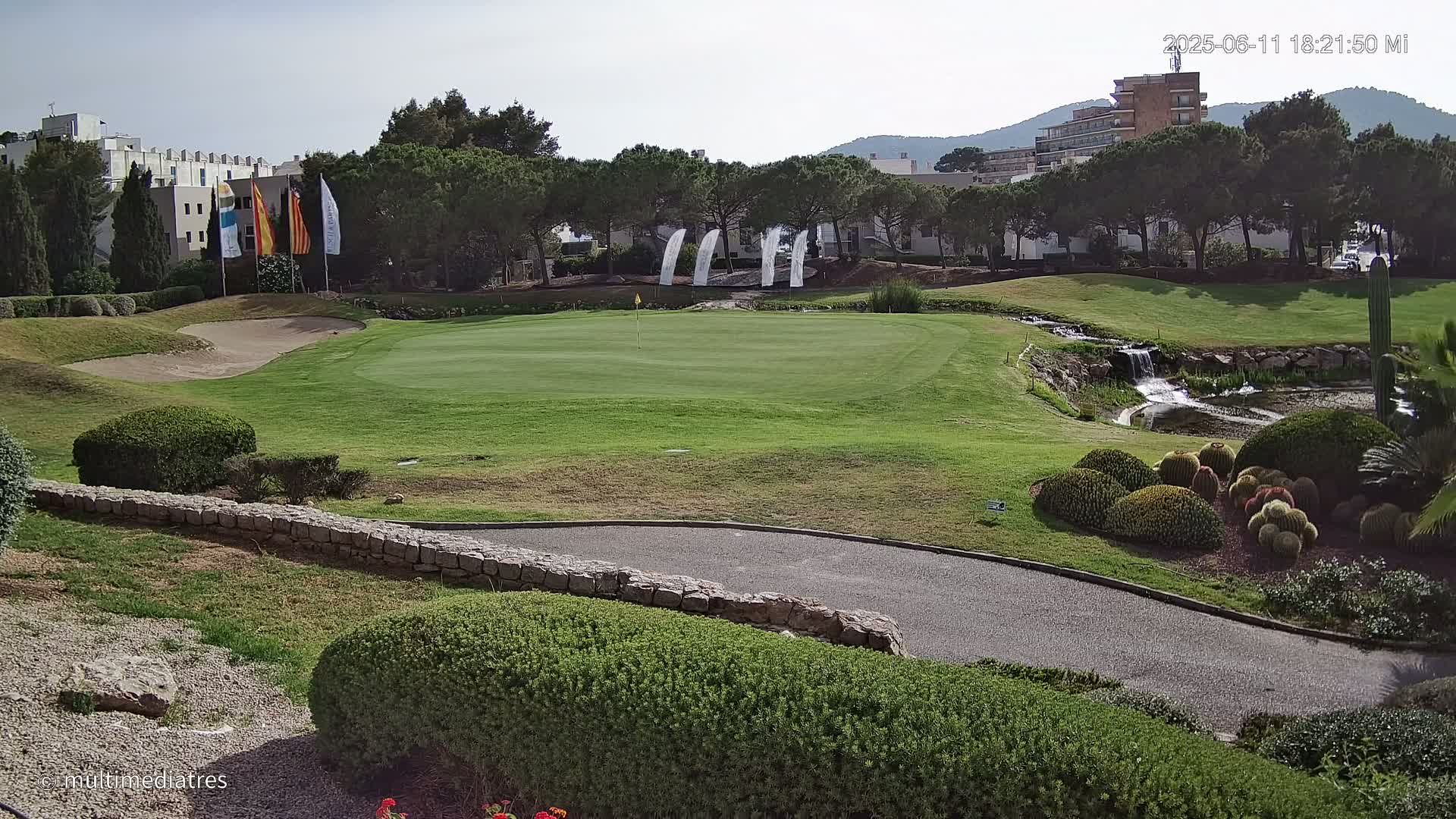 A golf course green with a sand trap, a small waterfall, and manicured landscaping under a mostly clear sky.
