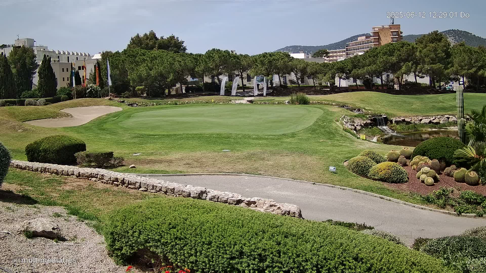 A golf course green with a sand trap, a small waterfall, and various shrubs and cacti, situated near buildings under a sunny sky.