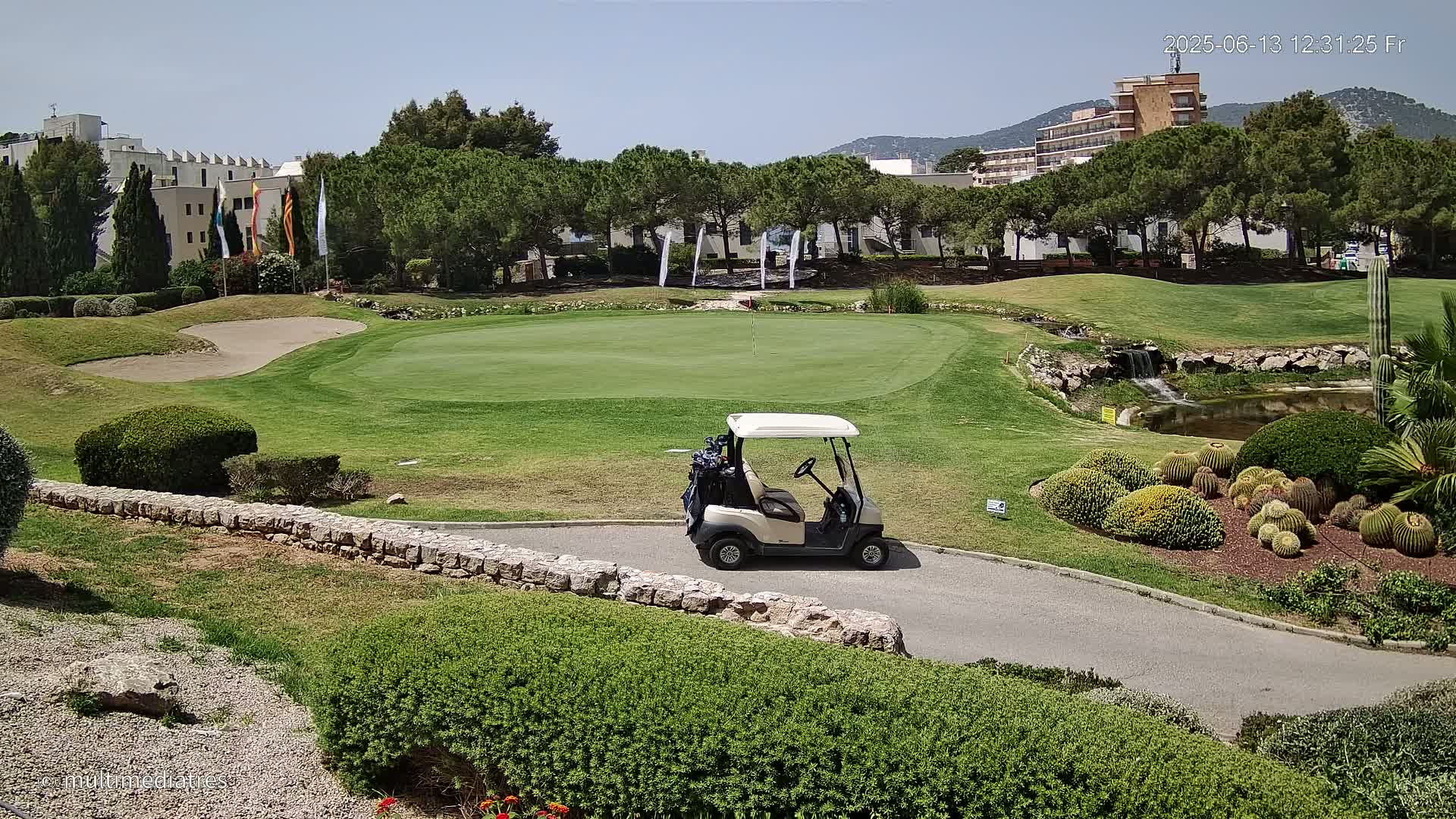 A golf cart sits on a paved path next to a putting green, with a small waterfall and various cacti visible in the sunny weather.