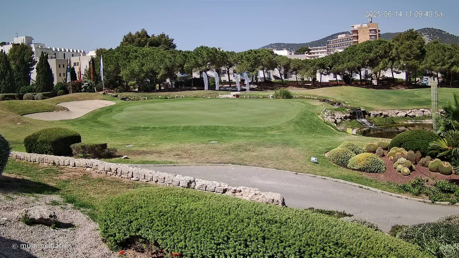 A golf course green with a sand trap, a small pond with a waterfall, and manicured landscaping, all under a sunny sky.
