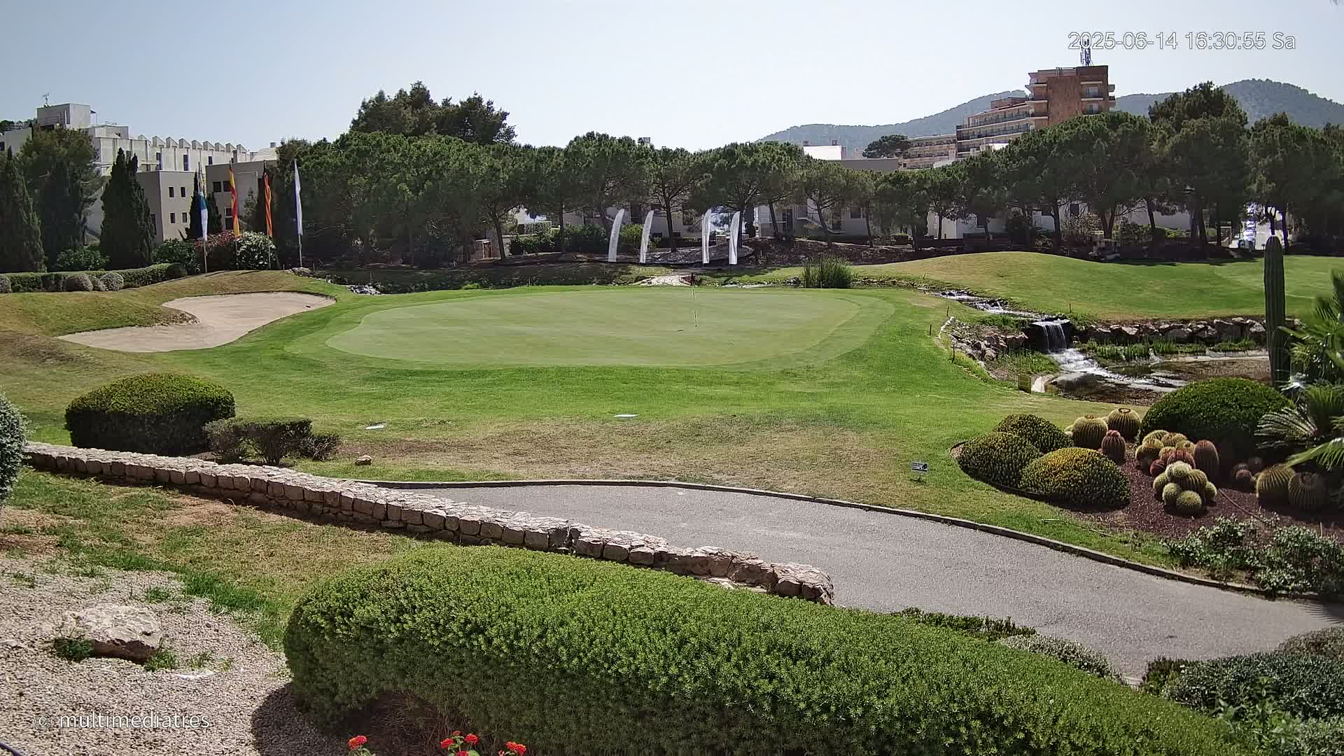 A sunny golf course green with a sand trap, a small waterfall, and manicured landscaping surrounds a low stone wall and paved pathway.