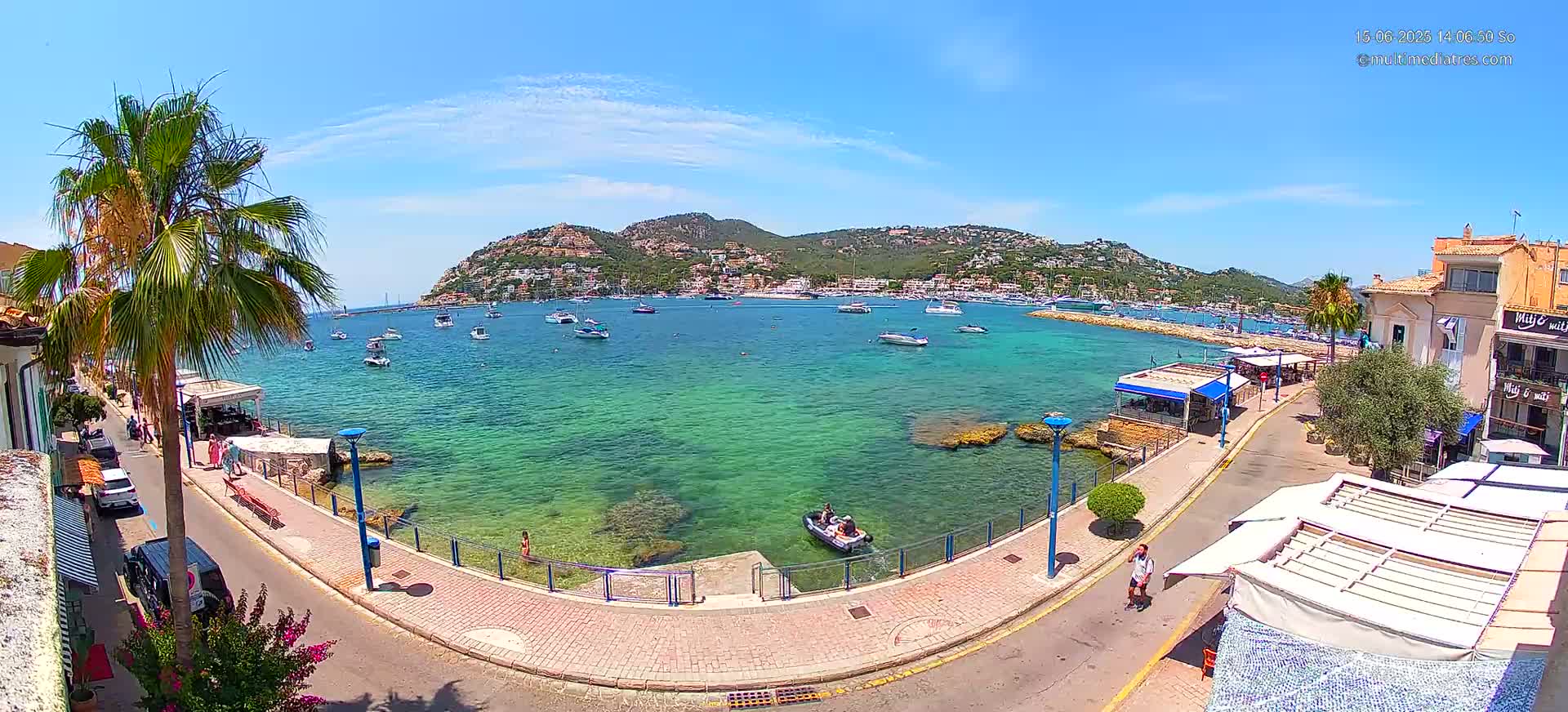 A clear, sunny day reveals a waterfront scene with calm, turquoise water filled with boats, a paved walkway lined with buildings and palm trees, and a small, rocky shoreline.