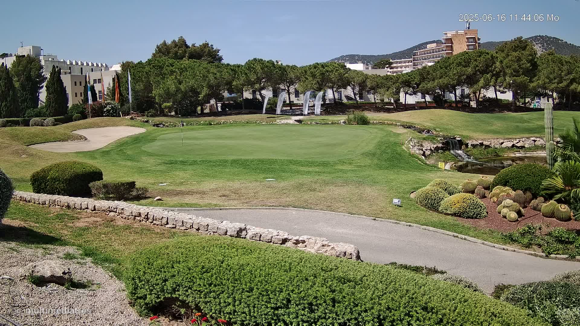 A putting green on a golf course is situated near a small waterfall and pond, with a building and trees in the background under a clear sunny sky.