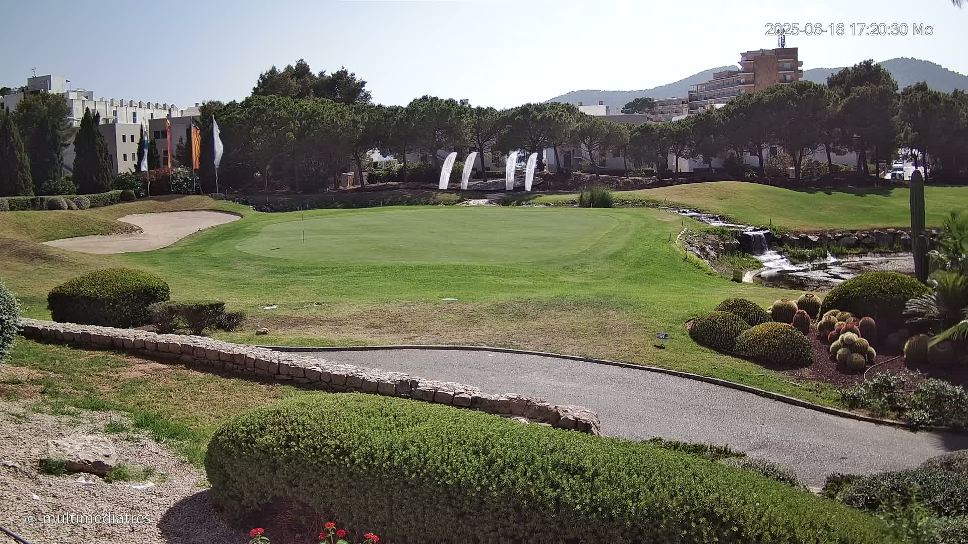 A golf course green with a small waterfall and several buildings in the background on a sunny day.